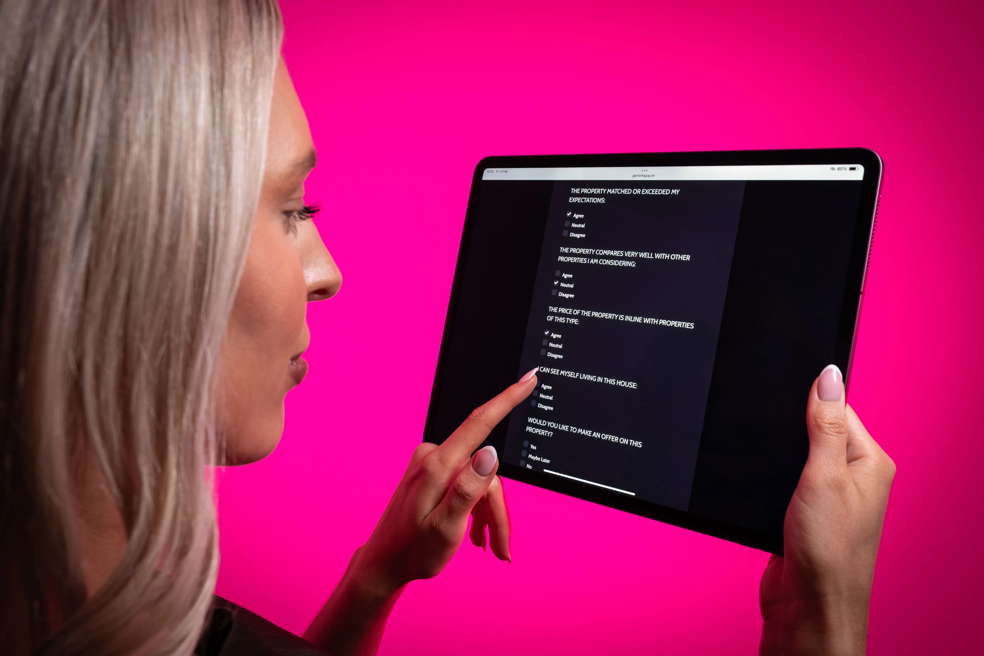 A person with long blonde hair uses a tablet displaying text on a dark screen. The background is a vibrant pink, creating a modern and focused atmosphere.