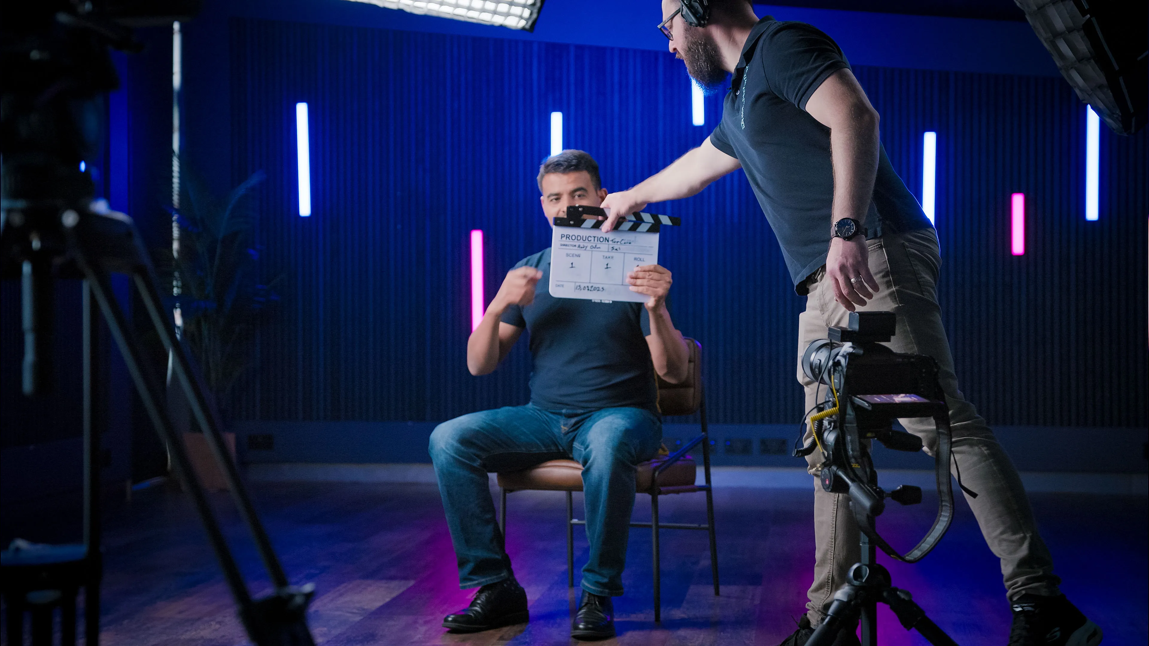 A man sits on a chair under studio lights, holding a film clapperboard. A crew member adjusts the clapperboard. The room has colourful neon lights, creating a professional and focused atmosphere.