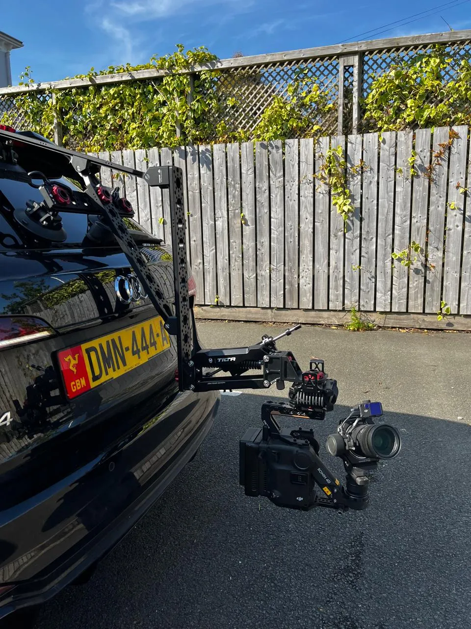 Tilta Hydra Alien arm and DJI Ronin 4D cinema camera mounted to the rear of a black vehicle with Isle of Man registration plates, showing the full rig from a three-quarter angle during a DotPerformance production