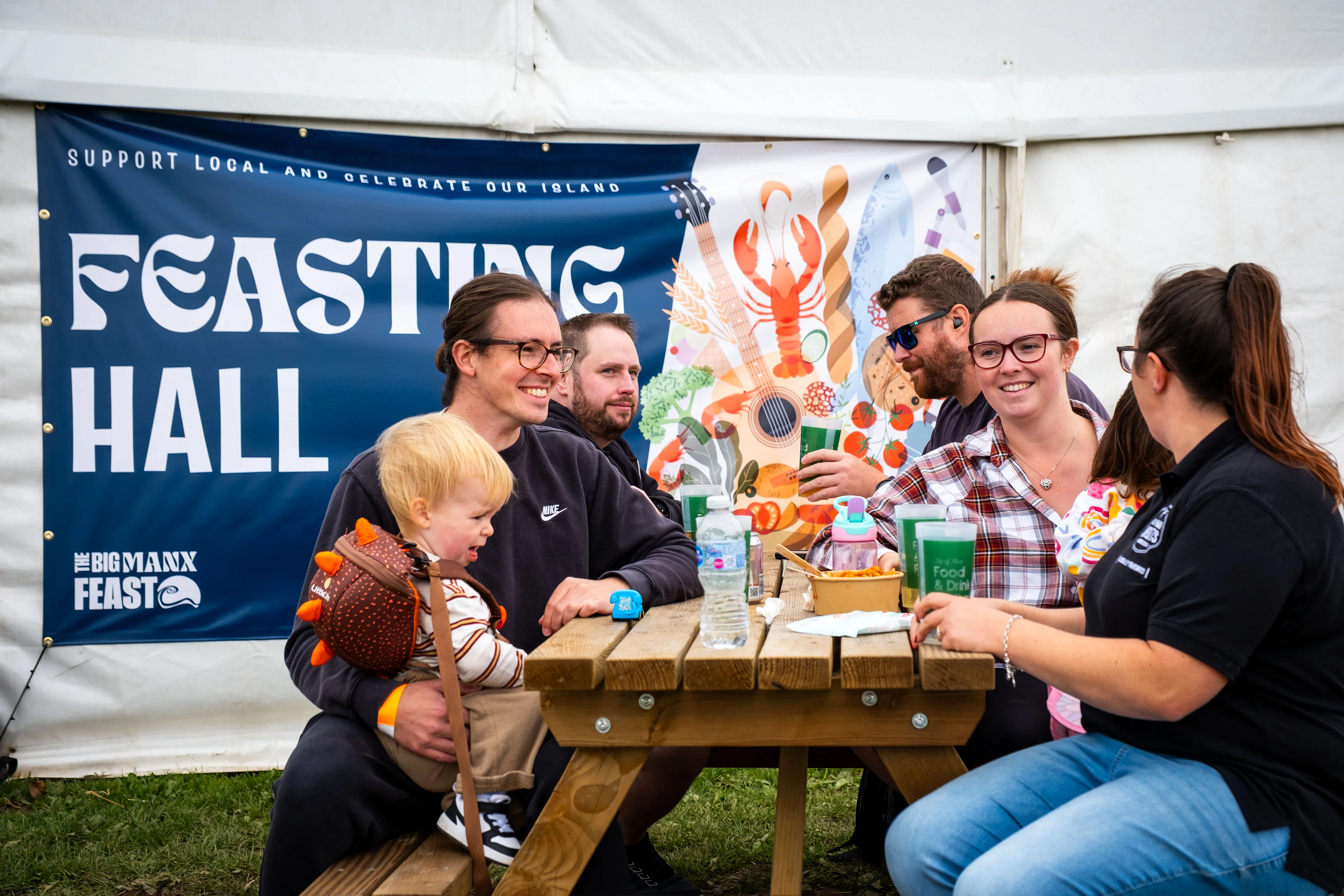 A group with a young child laughing around a picnic table outside the Feasting Hall at the Big Manx Feast