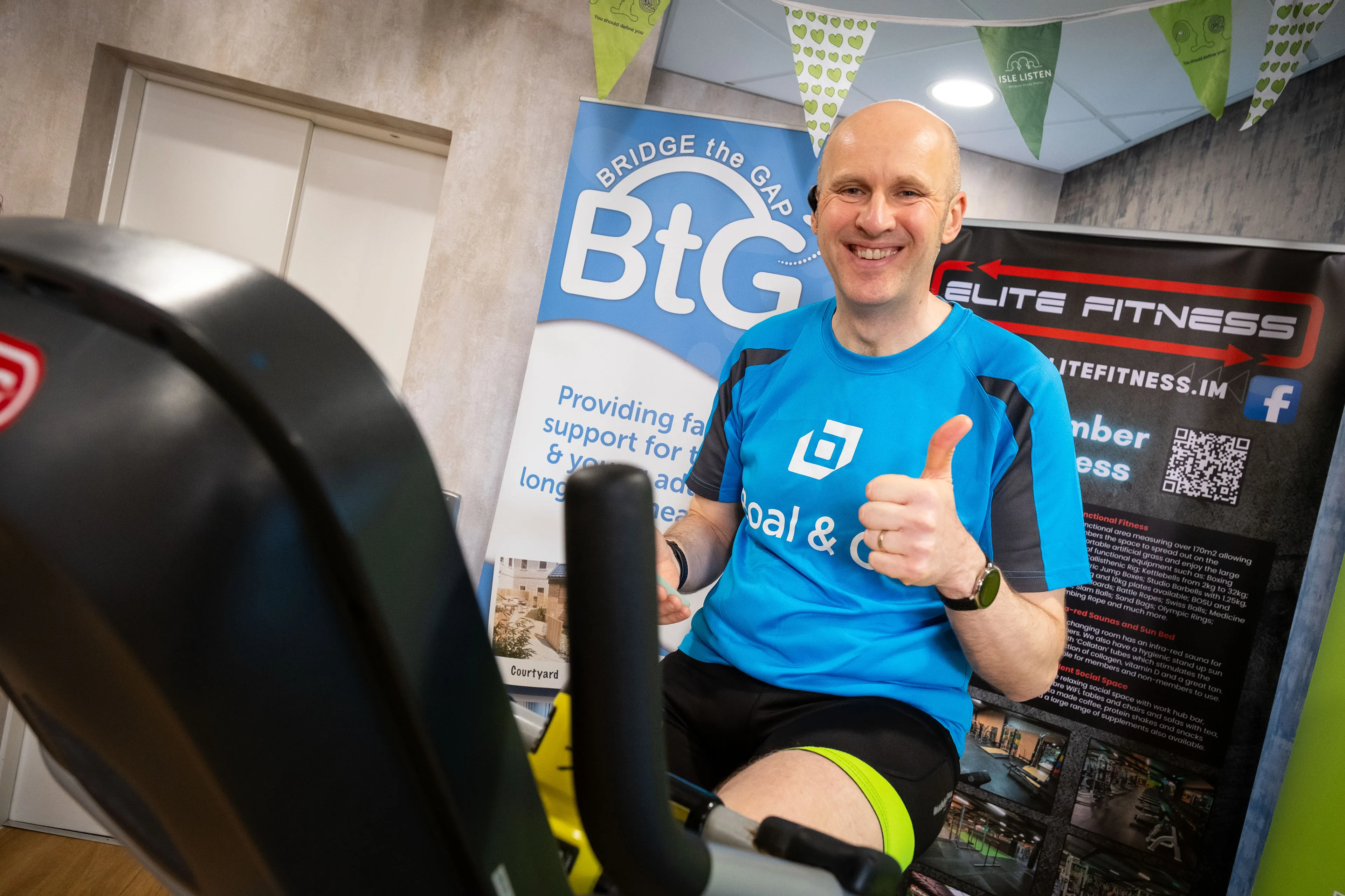 James Bassett on a static bike giving a thumbs up during the 12-hour charity cycling challenge on the Isle of Man, with a Bridge the Gap banner in the background
