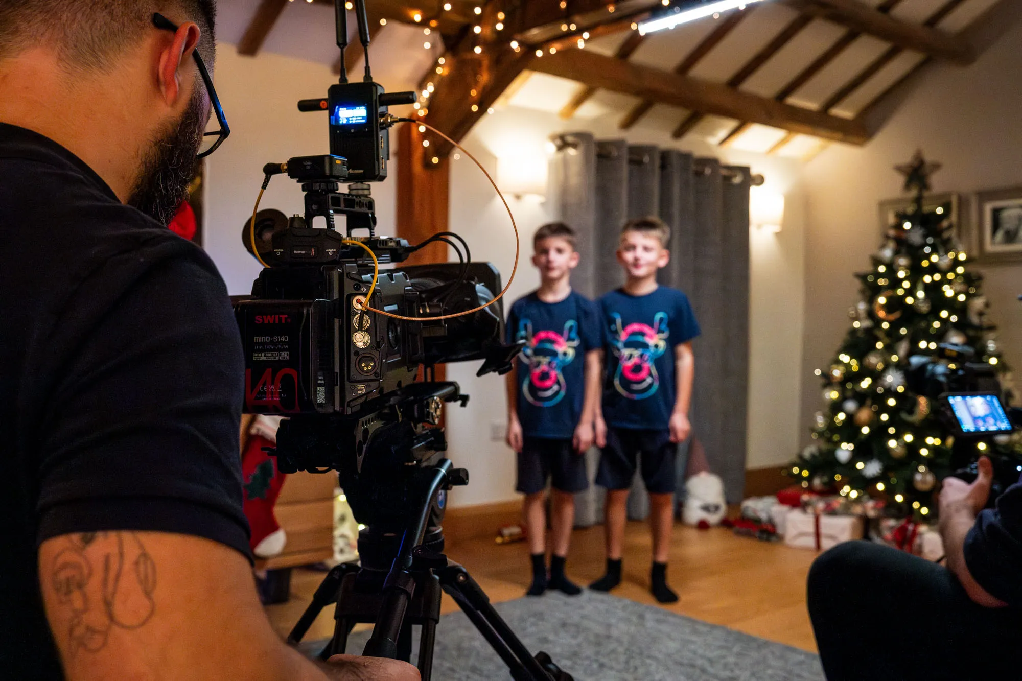 A filmmaker captures two boys standing in matching shirts beside a decorated Christmas tree. The festive room has soft lighting and wrapped gifts.