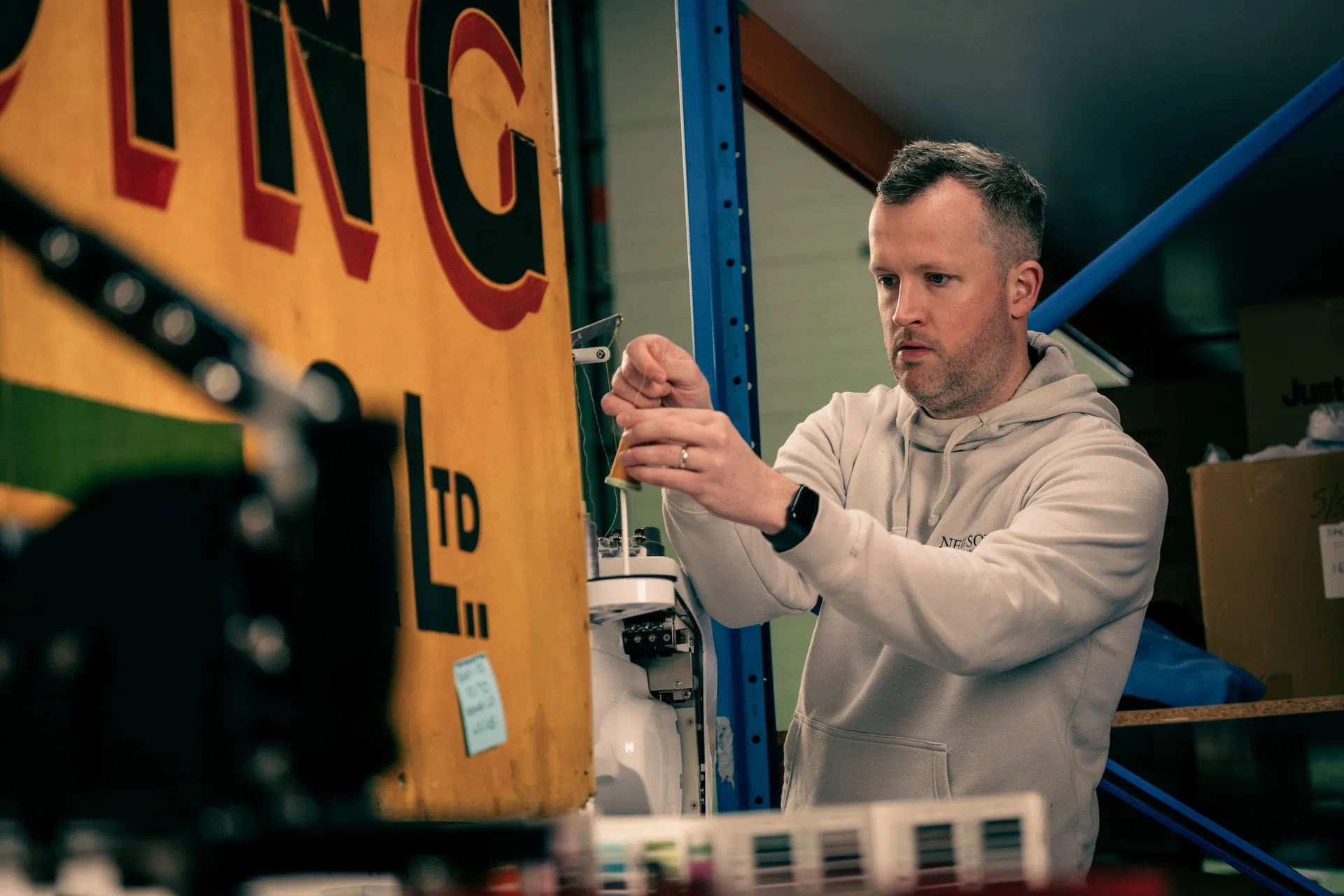 A man in a hoodie works intently with tools in a workshop. Surrounded by shelves, boxes, and machinery, he appears focused and precise.