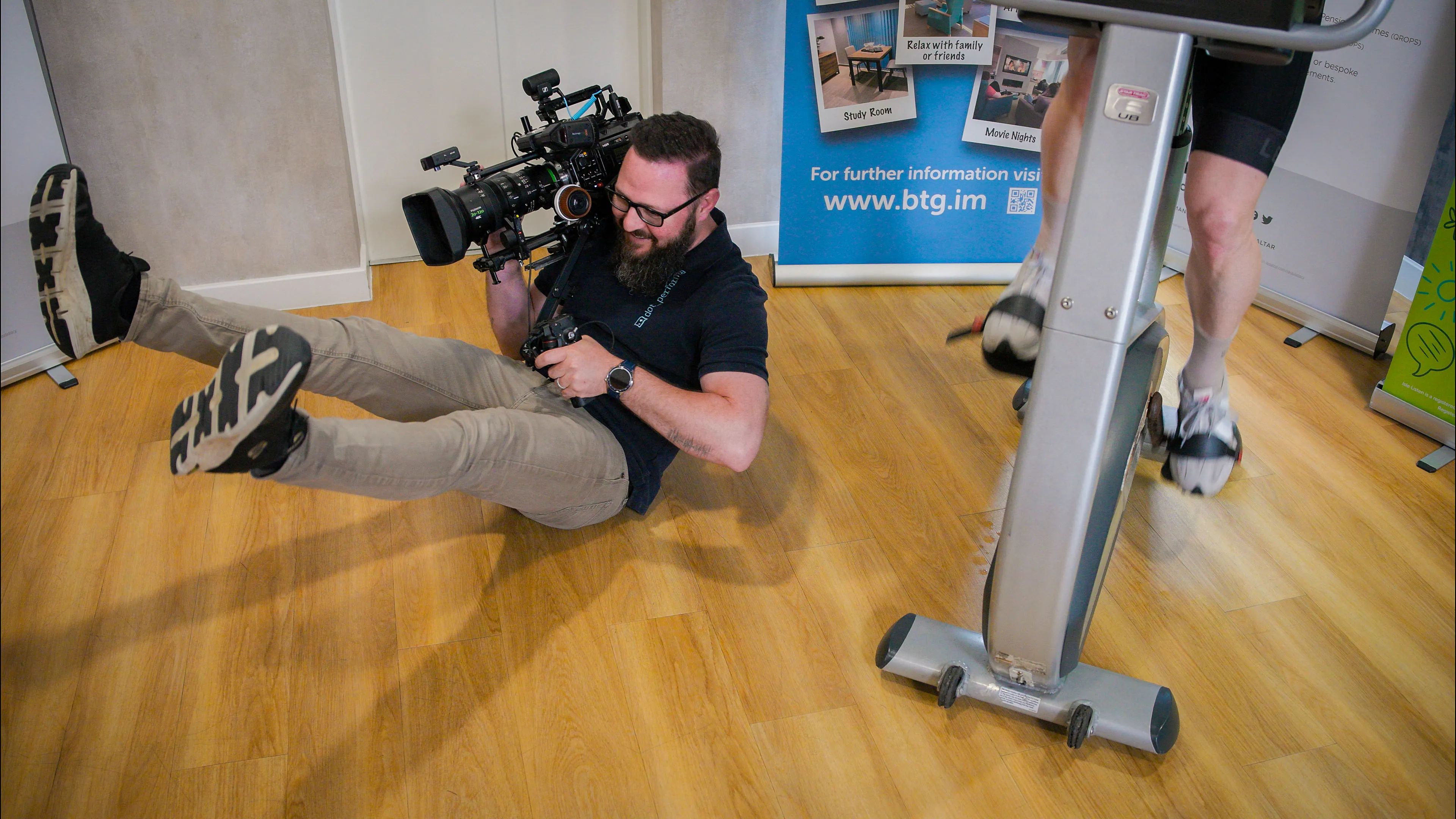 Nico van Loggerenberg from DotPerformance on the floor of the Boal & Co office with a Blackmagic URSA 12K Mini Pro, filming a rider on a static bike at low angle during the charity cycling challenge on the Isle of Man