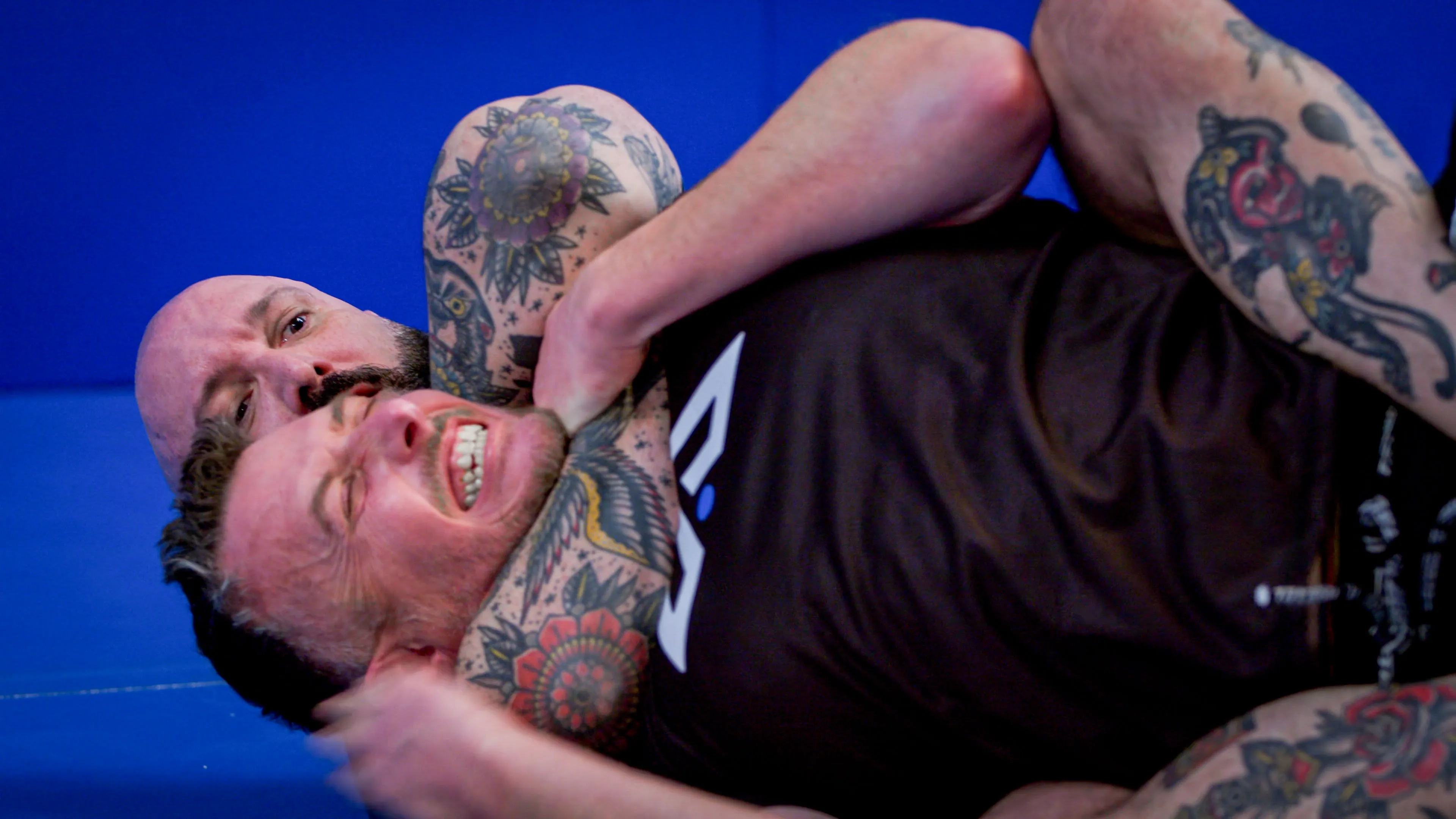 Two men engage in a grappling match on a blue mat. The man in front grimaces while the other applies a rear choke hold. Intense and competitive atmosphere.