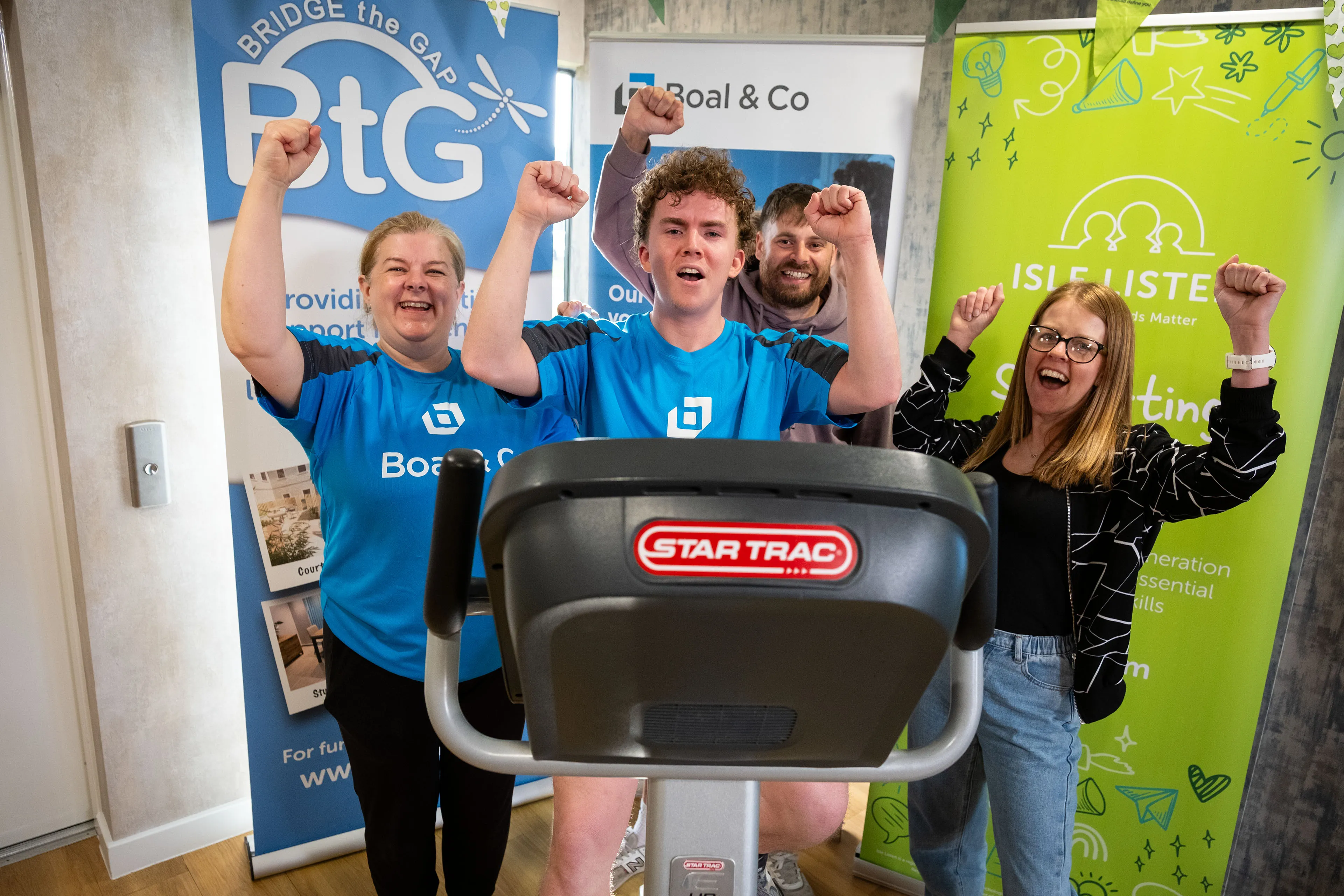 Christine Evans, Ed Butler, Andrew Cover and Vicky Mackie in branded cycling jerseys celebrating at the end of the 12-hour charity cycling challenge, with Bridge the Gap and Isle Listen banners visible in the background