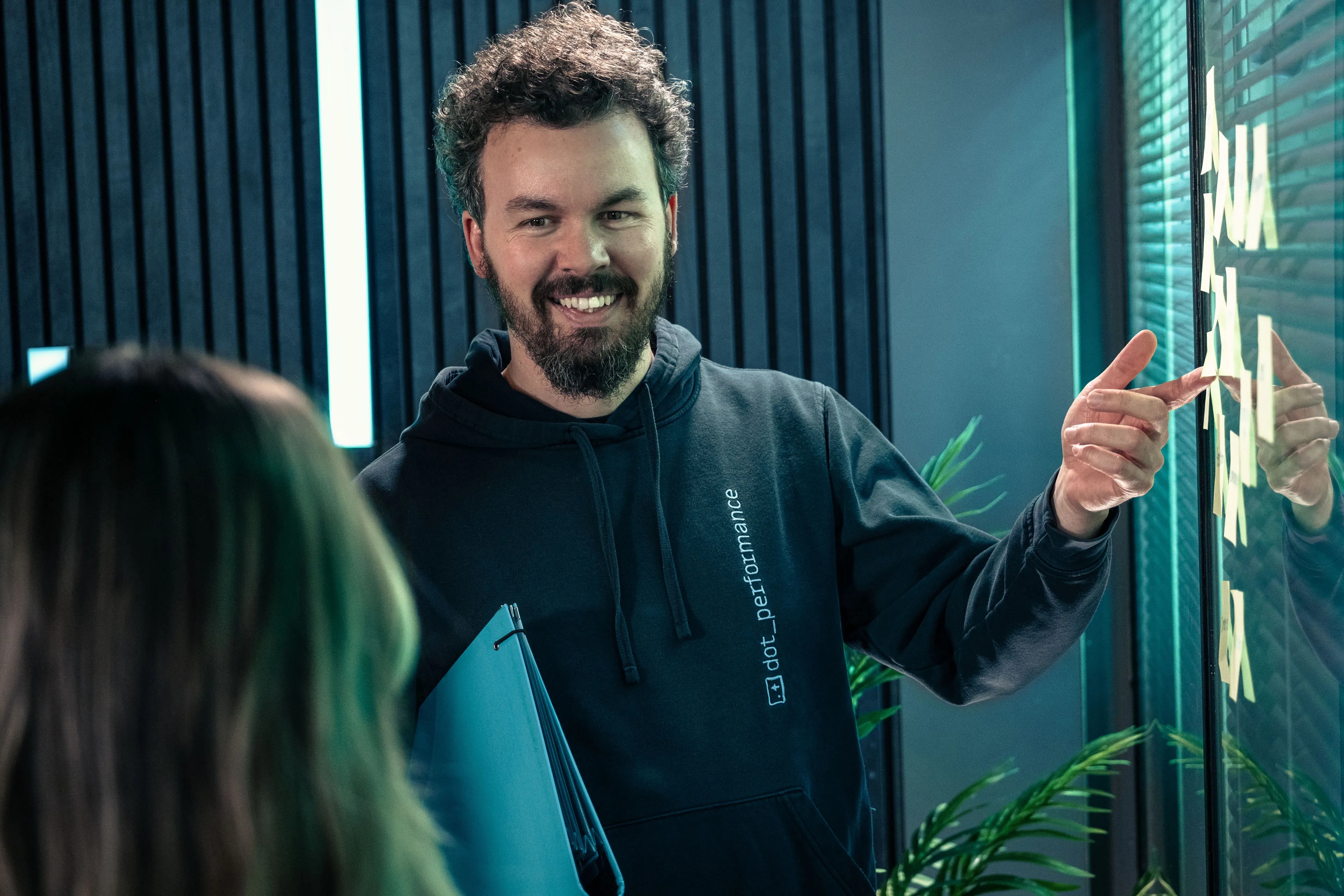 Adrian Crellin presenting UX wireframe notes on a glass planning wall at DotPerformance studios on the Isle of Man, wearing a DotPerformance hoodie