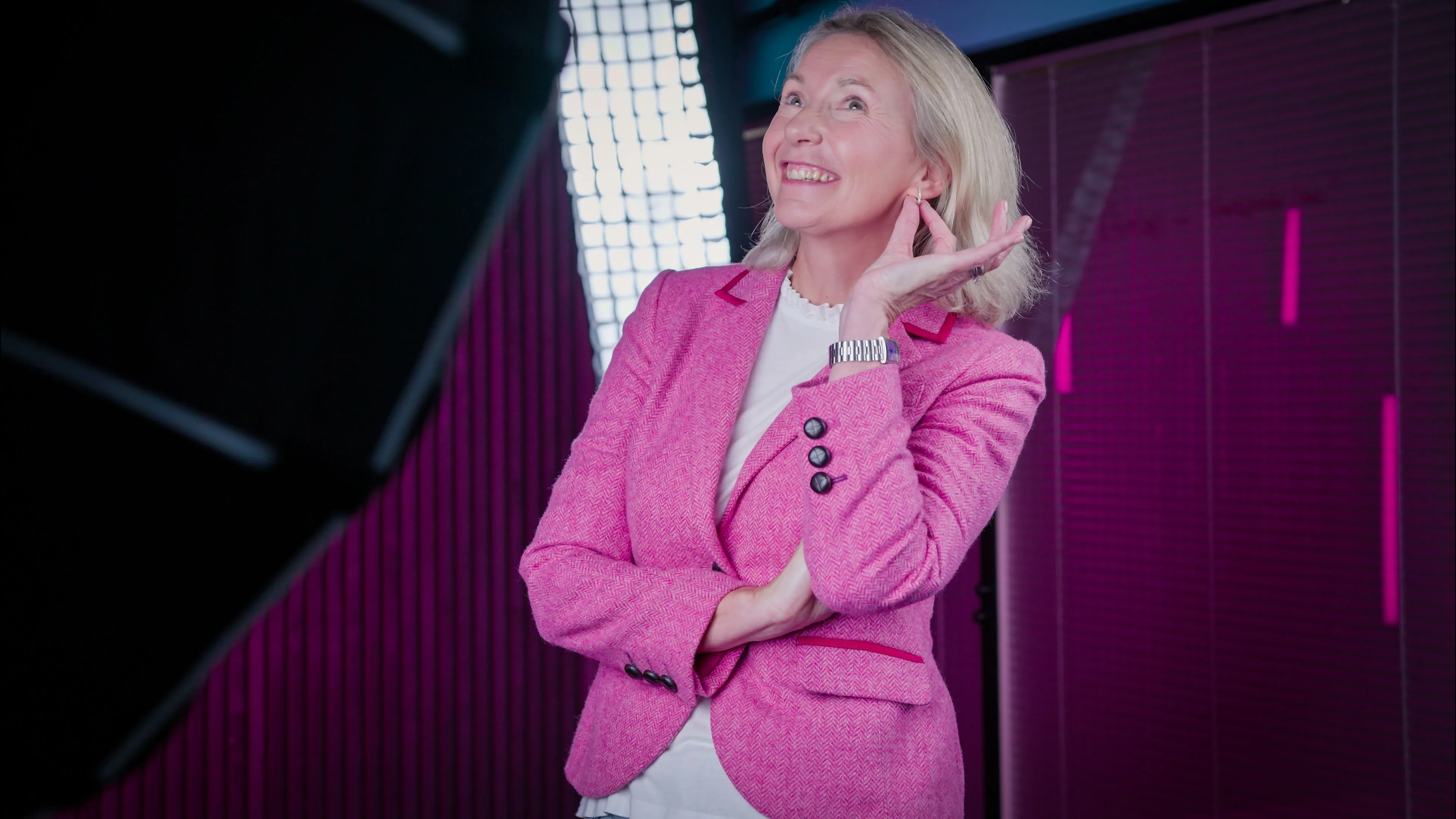 A woman in a bright pink blazer poses confidently with a smile, hand on her ear. She's in a modern studio with vibrant purple lighting, exuding a cheerful and professional tone.