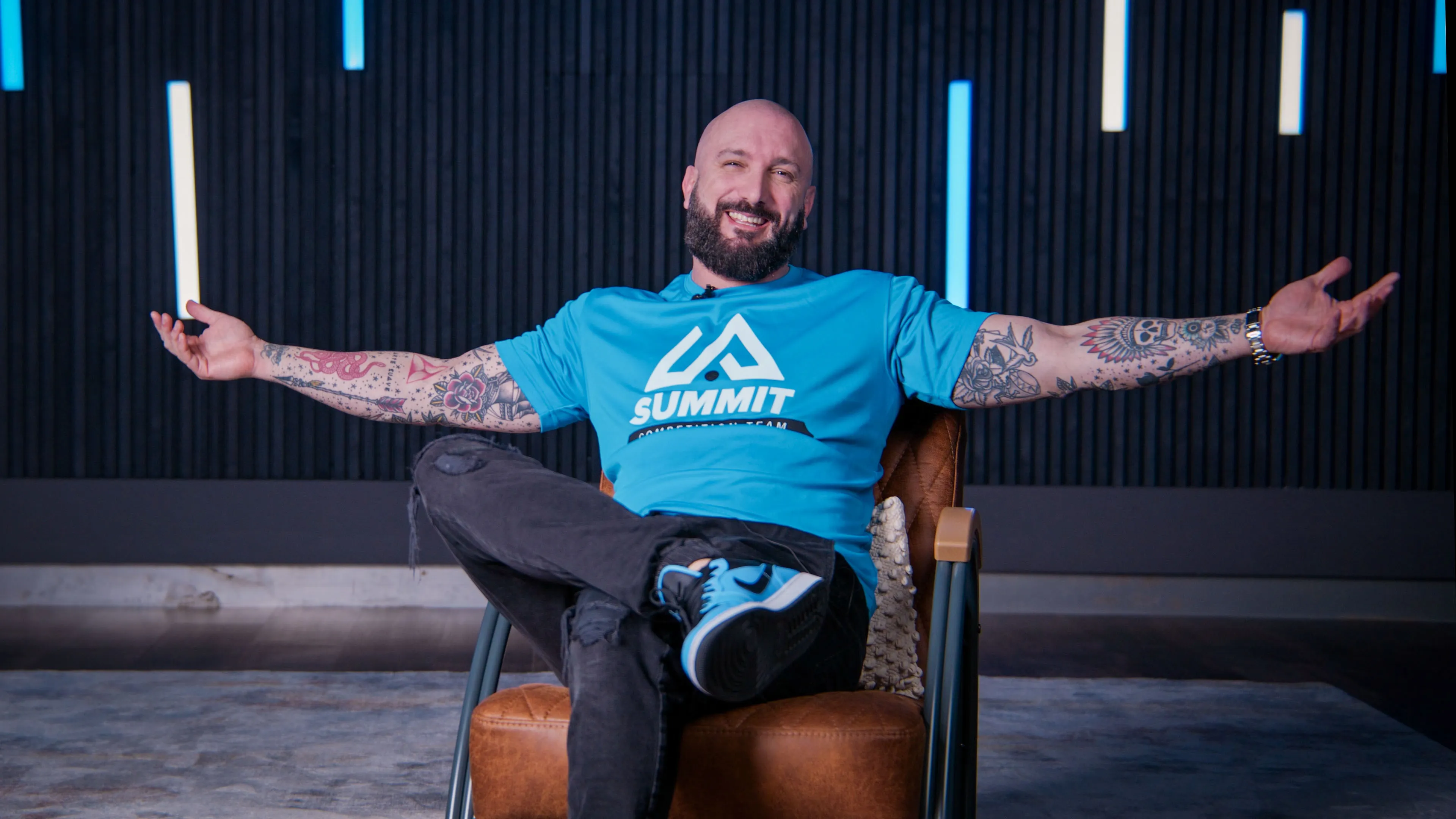 Tattooed man smiling with outstretched arms, wearing a blue "Summit" t-shirt. Seated on a brown chair in a modern room with vertical light strips.