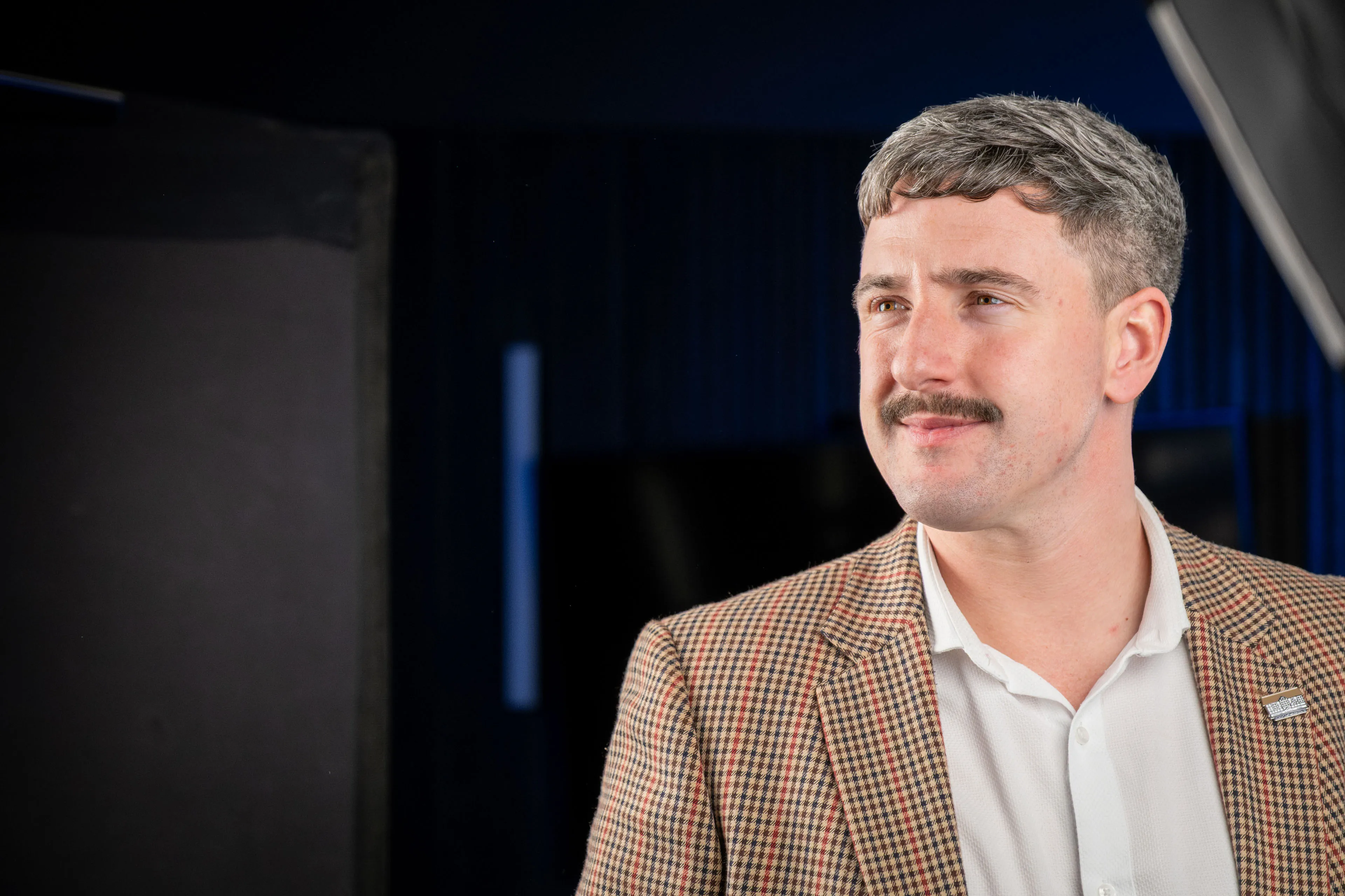 An ECAP employee with a moustache and tweed jacket smiling for his headshot against a dark studio backdrop with blue accent lighting