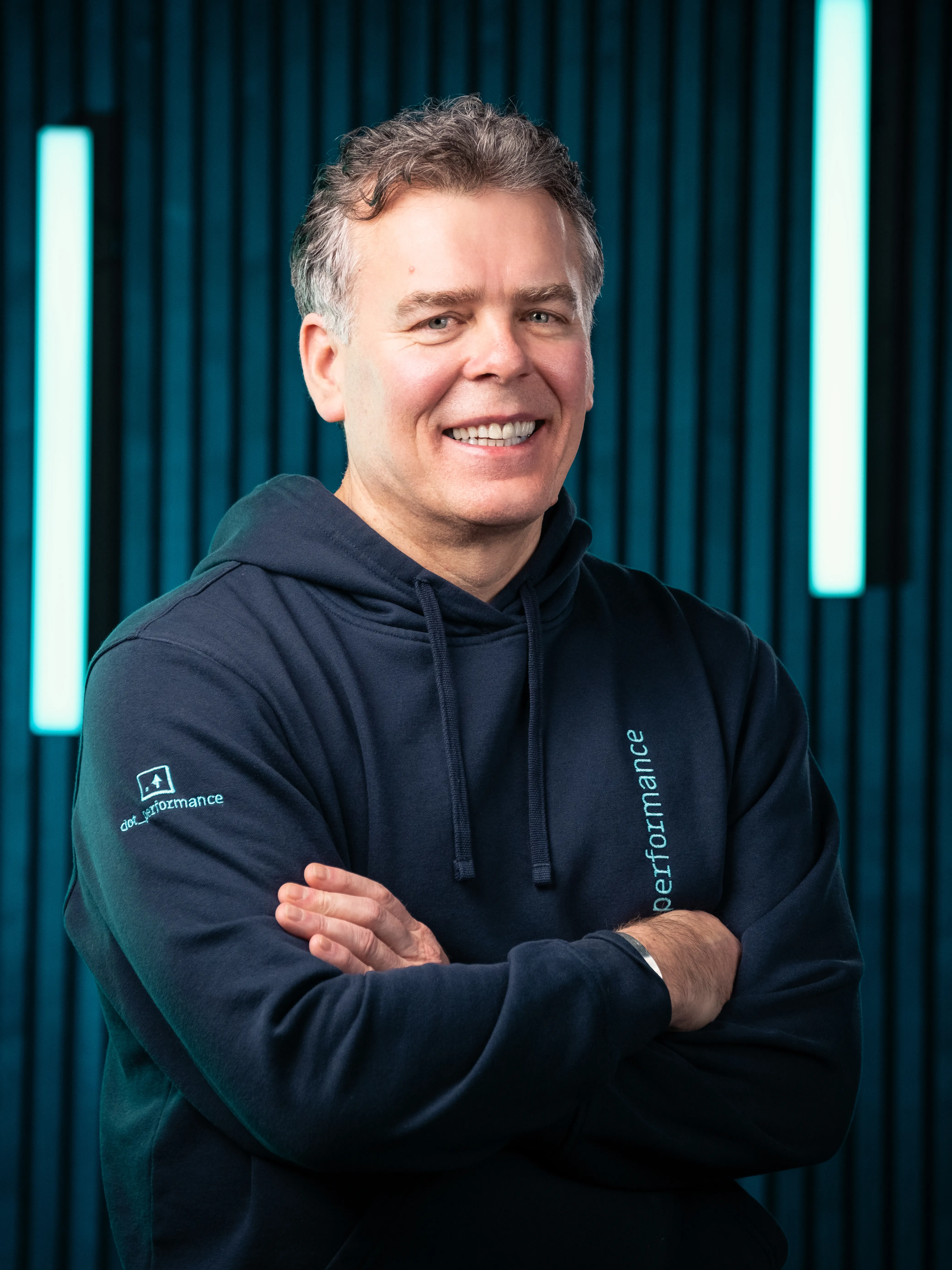 Jason, branding specialist at Dot Performance, smiling with crossed arms as he poses for headshot wearing branded hoody, against dark background with blue neon lights.