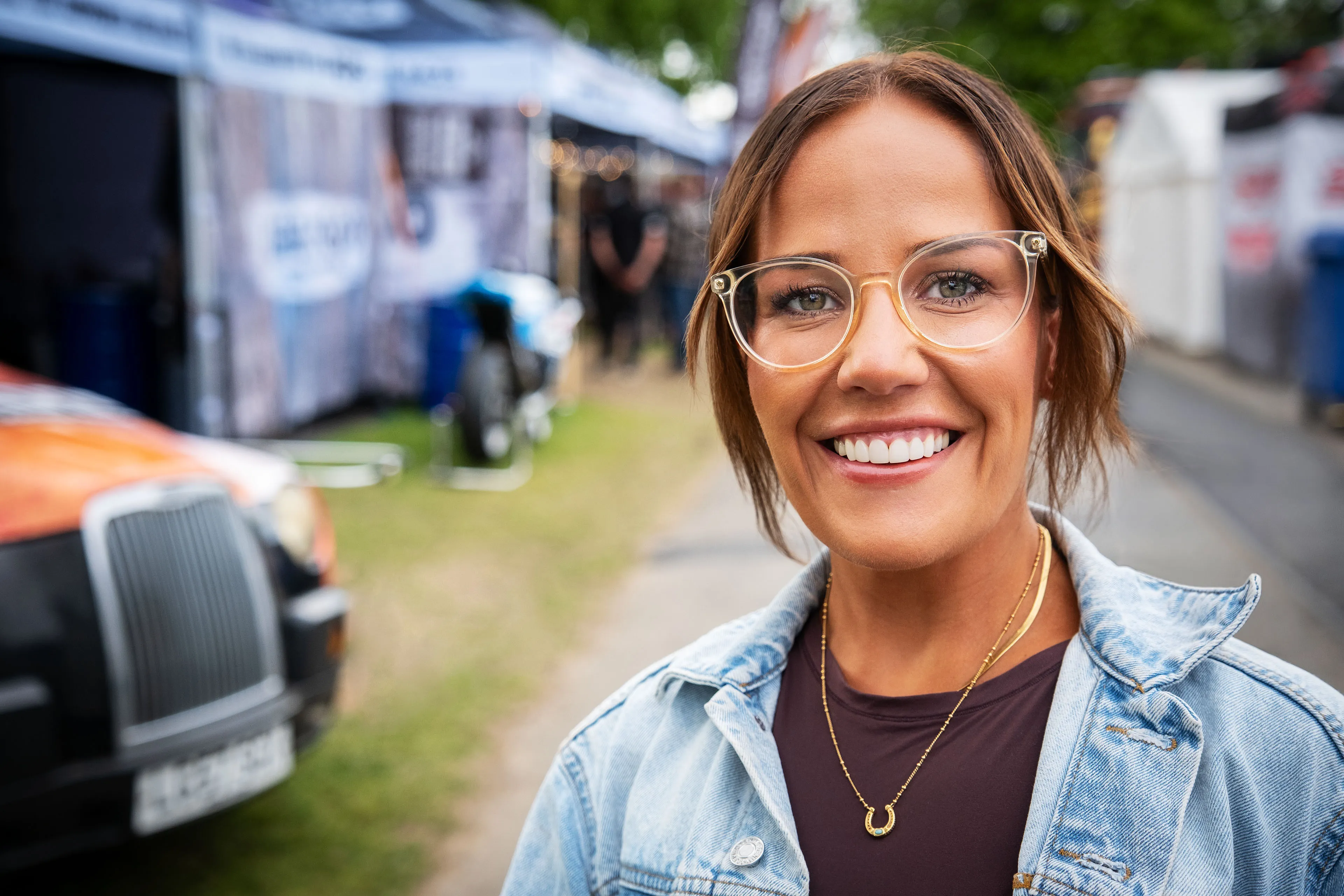 Nell Ransom from Leeds, living in Texas, an Isle of Man TT Marshal, smiling outdoors for Behind the Tabard TT Marshals Series highlighting marshal stories, in a denim jacket, and perspex glasses