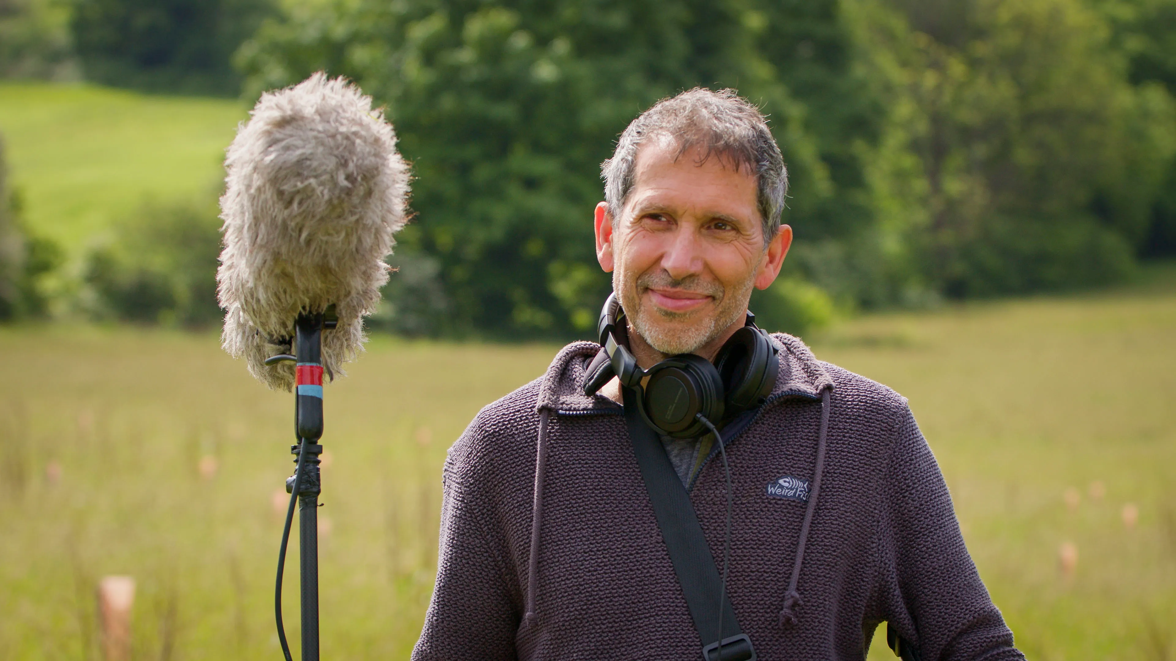 Man standing in a grassy field with a microphone on a stand. He wears headphones around his neck, conveying a relaxed and focused tone.