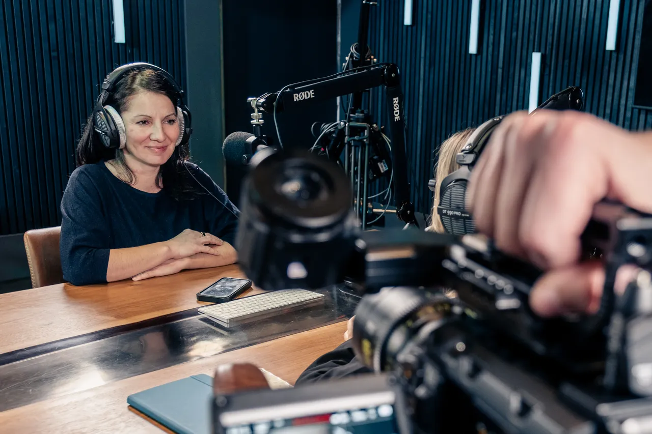 Ralu Matei Orton, DotPerformance's Operations Director, wearing headphones sat with a microphone in the DotPerformance podcast and recording studio on the Isle of Man, with an URSA Mini Pro 12K camera and Rode microphone visible in the foreground