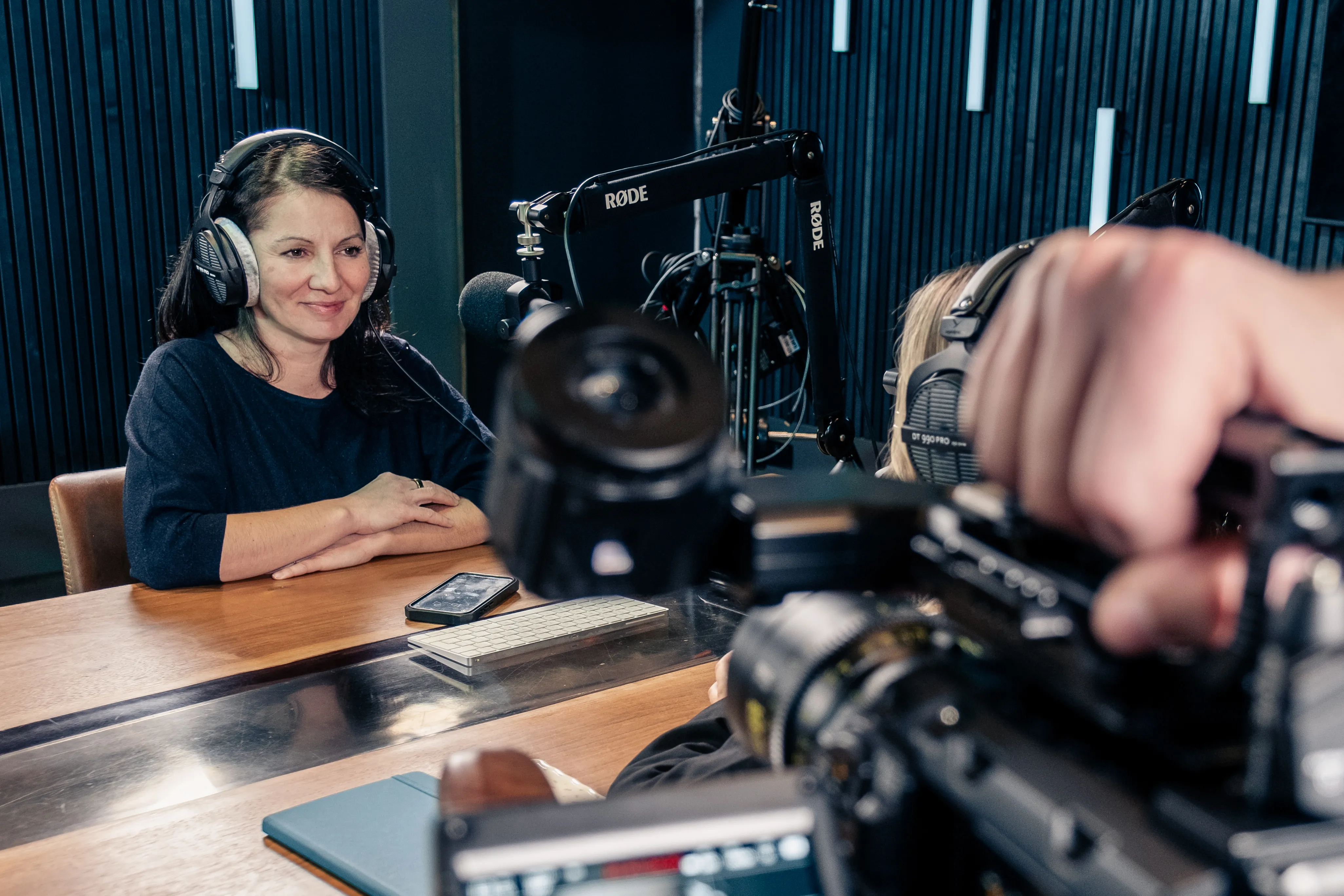 Ralu Matei Orton, DotPerformance's Operations Director, wearing headphones sat with a microphone in the DotPerformance podcast and recording studio on the Isle of Man, with an URSA Mini Pro 12K camera and Rode microphone visible in the foreground