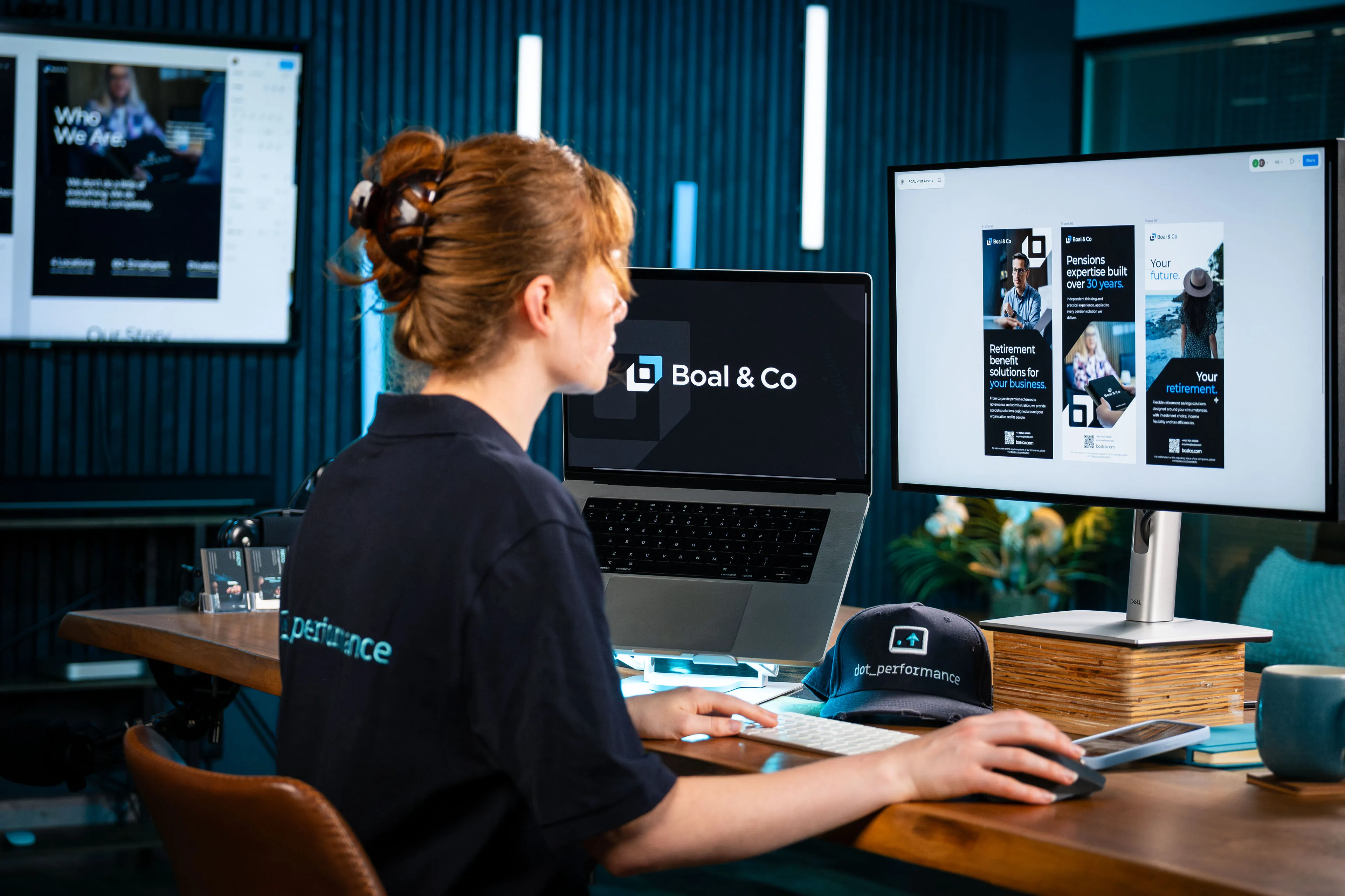 Ella Garvey sits at a desk in a DotPerformance polo shirt, working on Boal & Co print assets in Figma, including "Pensions expertise built over 30 years", beside a laptop showing the Boal & Co logo, at the DotPerformance office.