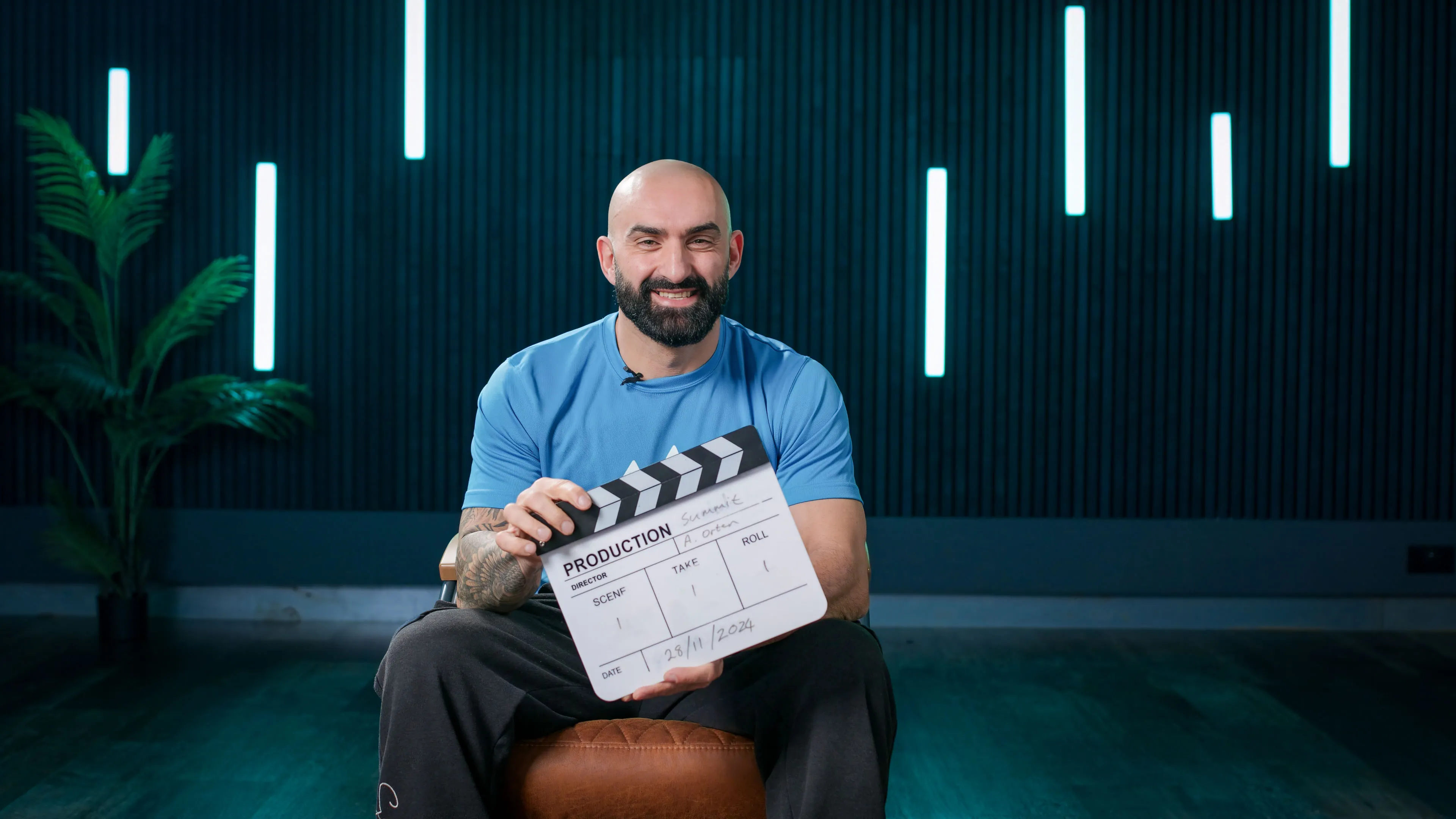 A smiling man with a beard, wearing a blue shirt, holds a clapperboard. He sits on a stool against a dark, modern background with vertical lights and a plant.