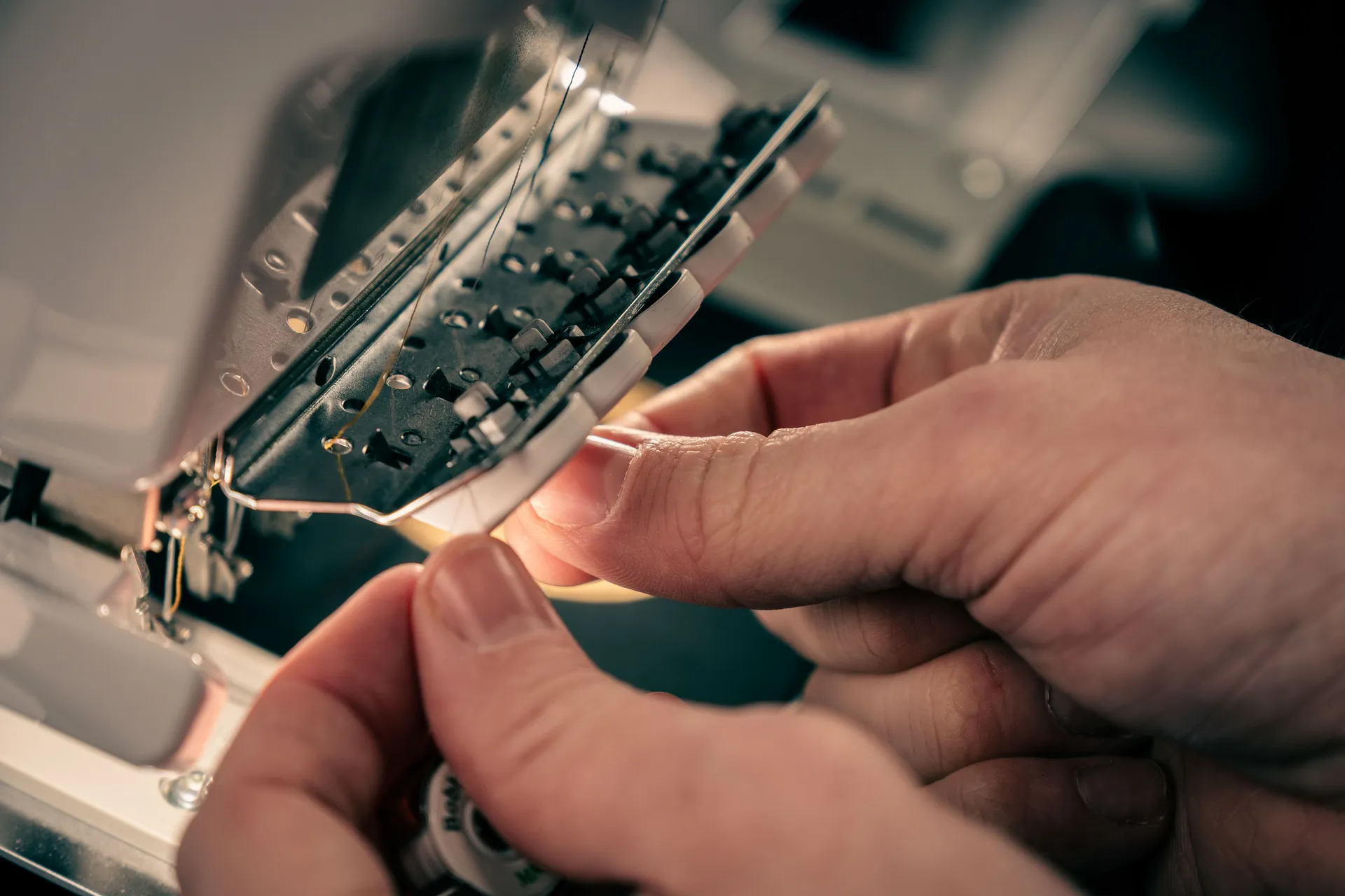 Close-up of hands meticulously threading a needle on a sewing machine, capturing a careful and focused moment with a dimly lit background.