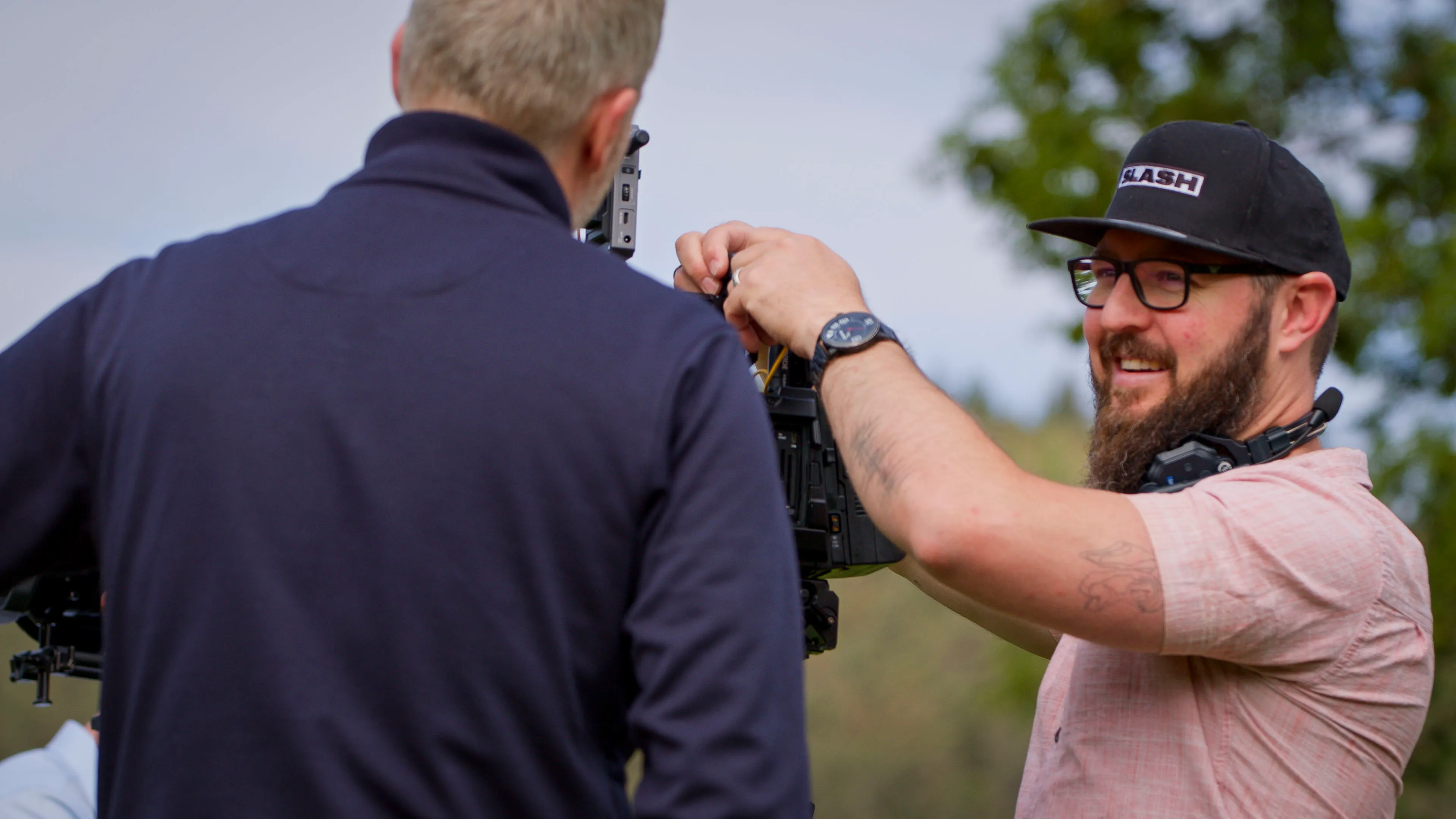 A bearded man in glasses and a cap adjusts a camera on a rig outdoors, conversing with a colleague in a dark jacket. The setting is relaxed and collaborative.