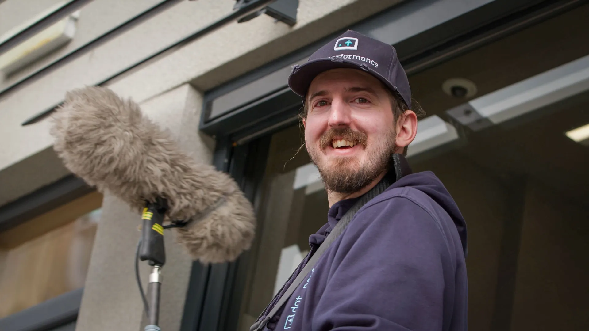 A man wearing a cap and hoodie smiles warmly while holding a boom microphone outside a building. The scene conveys a friendly, professional vibe.