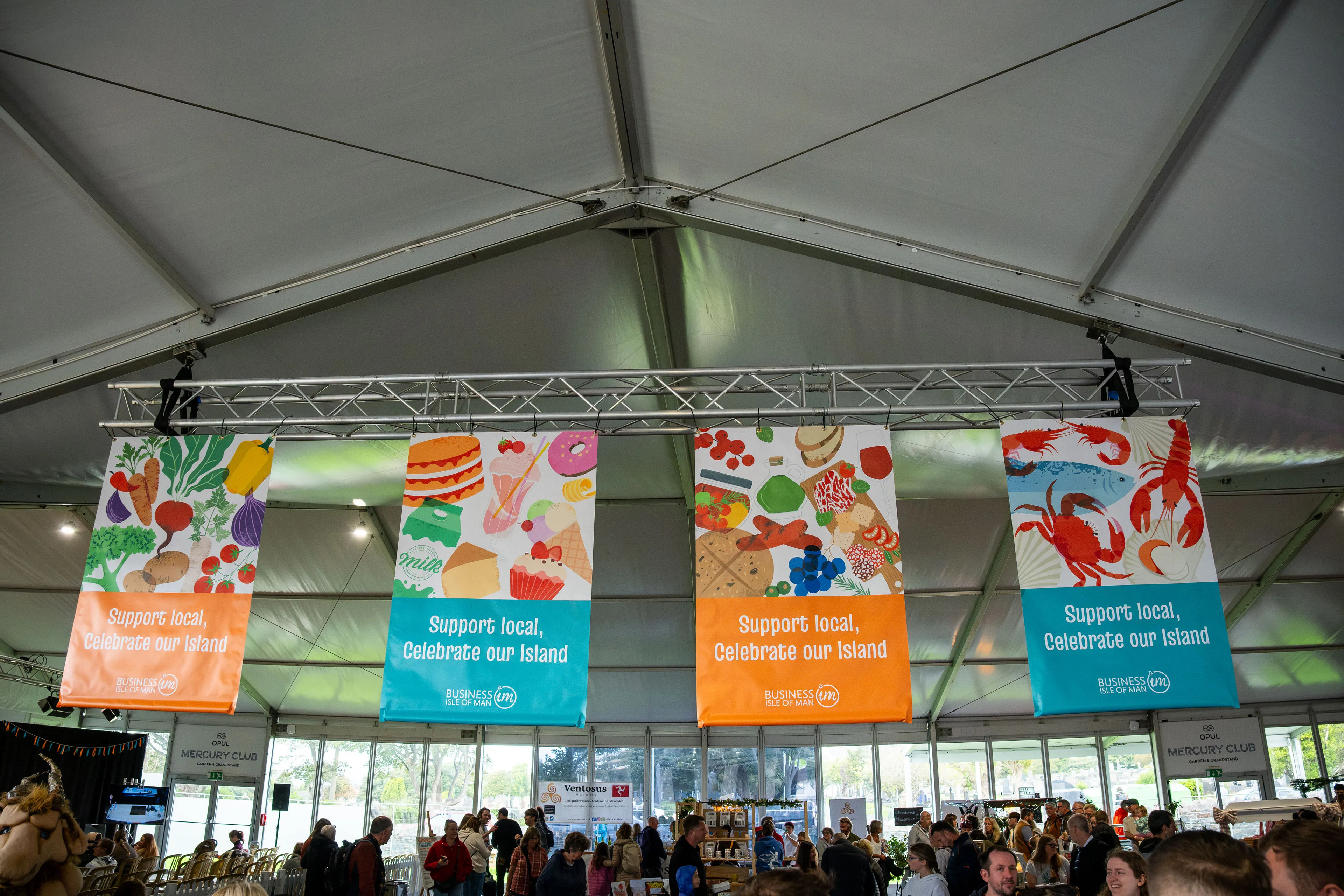Four "Support local, Celebrate our Island" banners with illustrated produce hanging in the Manx Produce Market hall at the Big Manx Feast