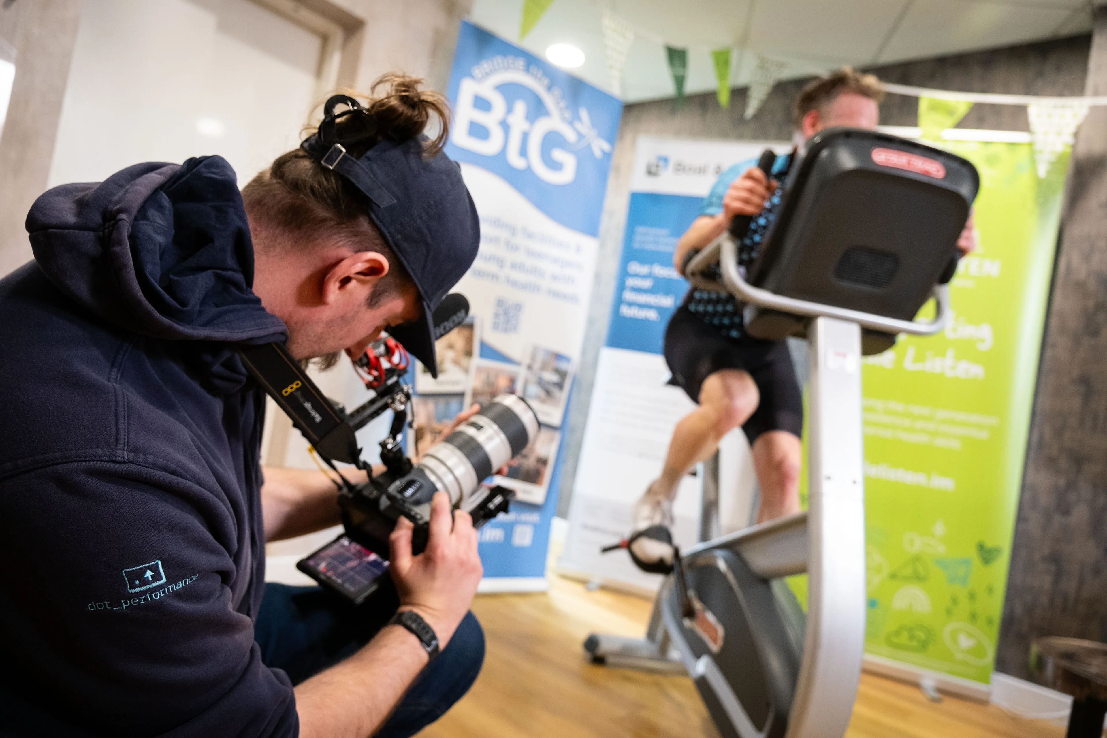 Will Oates from DotPerformance crouching to film John Beaty from DotPerformance using a Blackmagic Pocket 6K camera rig during the Boal & Co charity cycling challenge, with Bridge the Gap and Isle Listen banners visible
