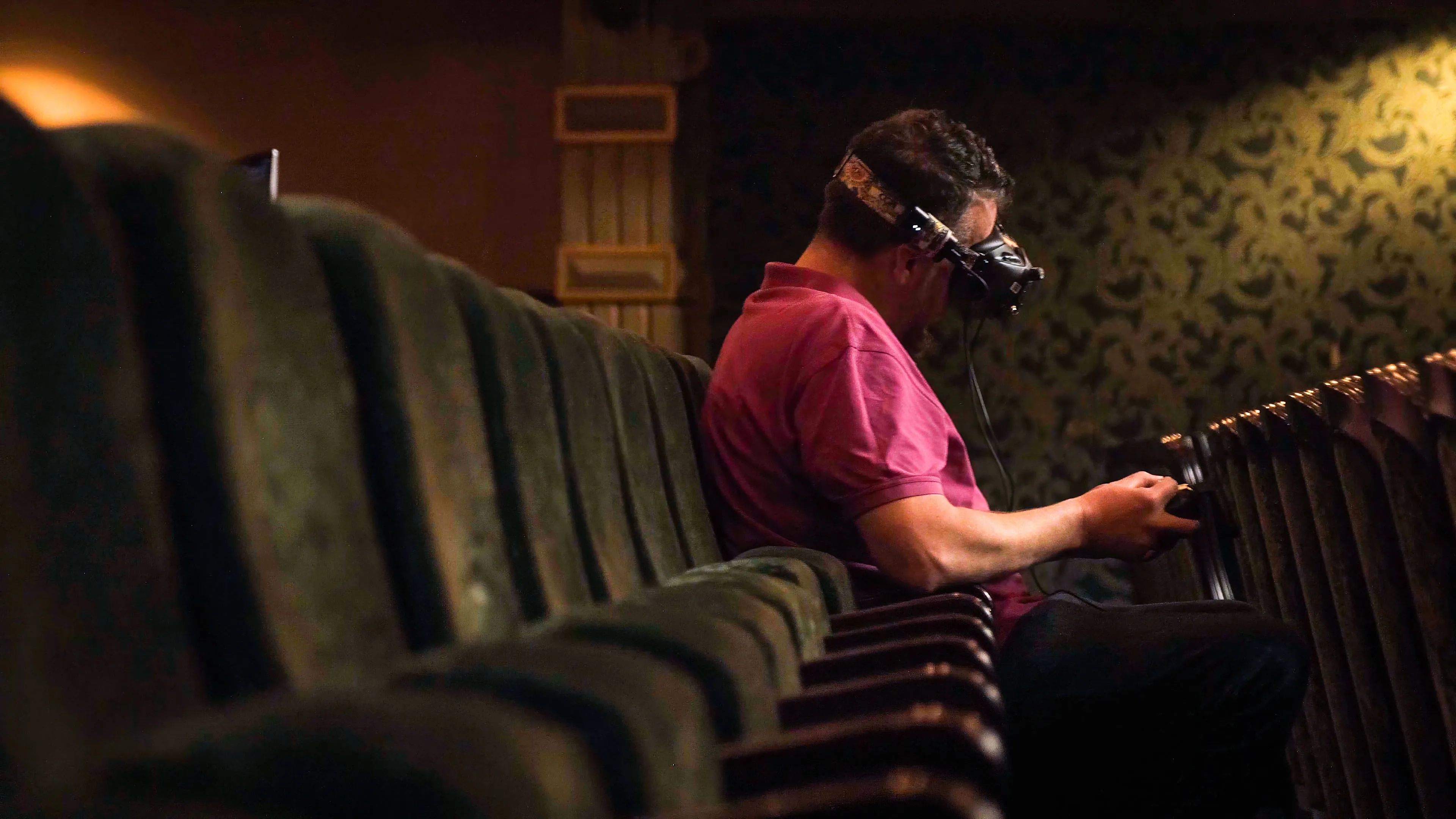 A person in a theater seat wears FPV Drone goggles, holding a remote, surrounded by empty rows. The lighting is dim and the mood is immersive and tech-focused.
