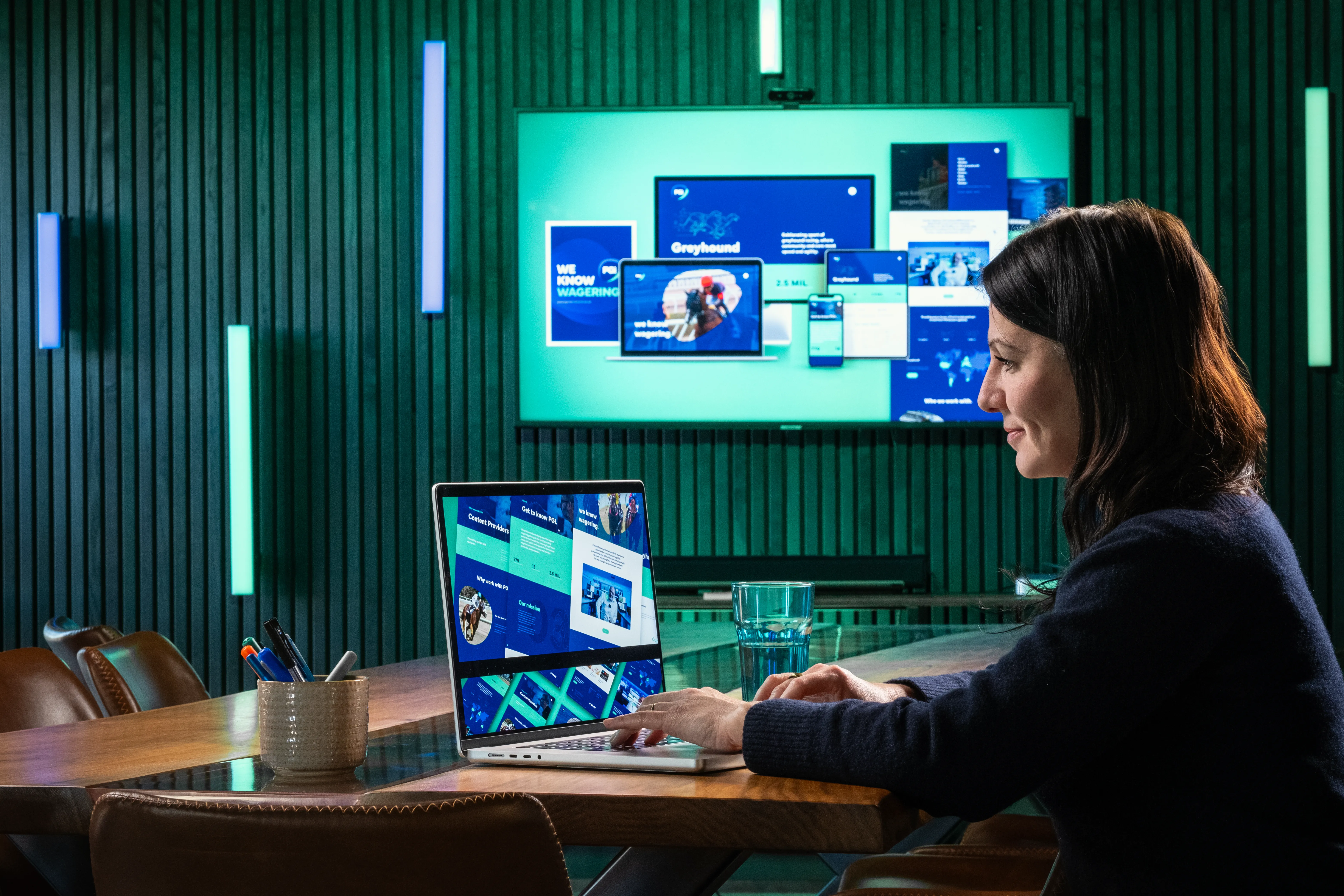 Raluca at a laptop in the Dot Performance studio, with PGI branding work shown on screens around her