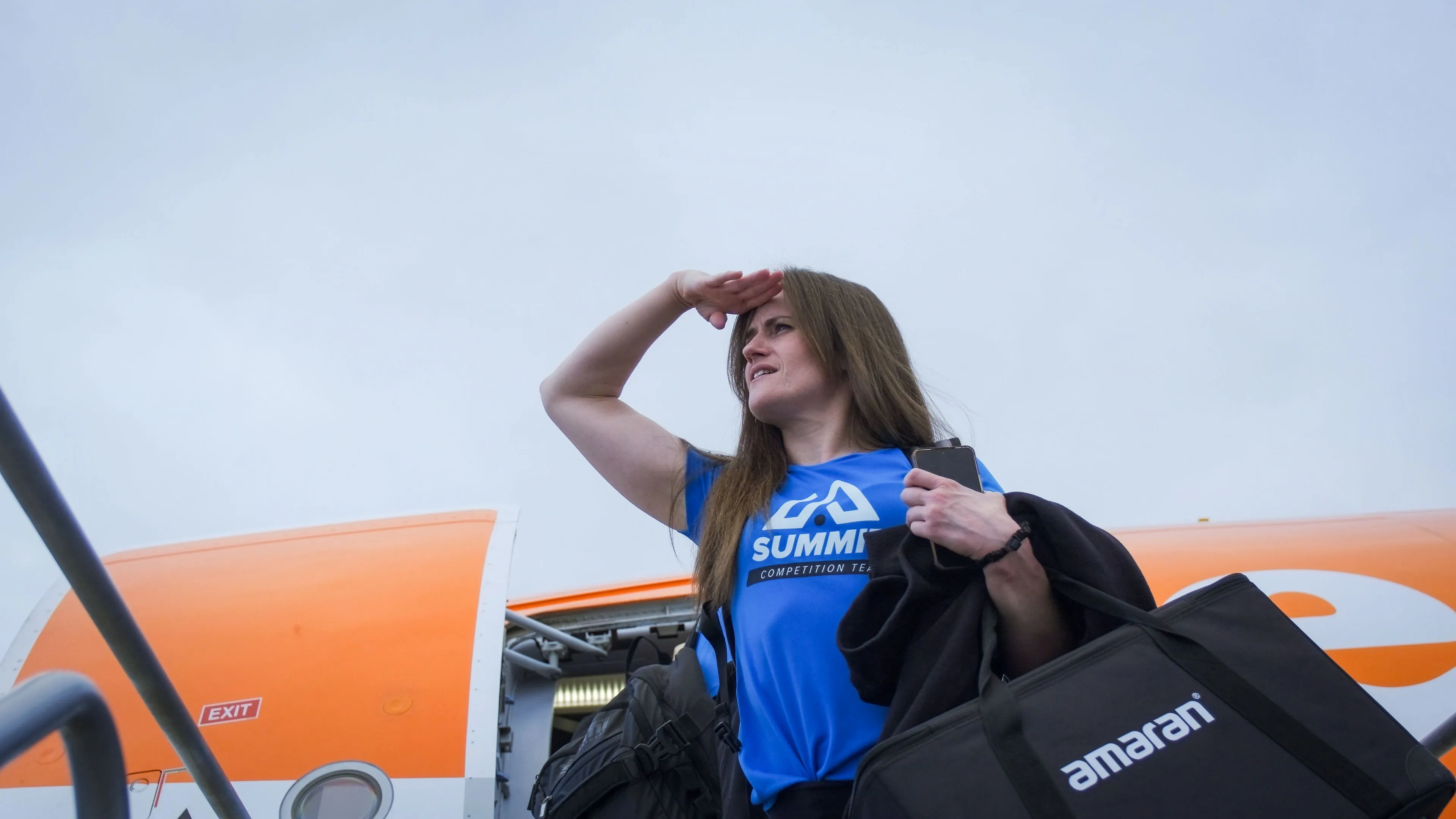Woman in a blue "Summit" shirt stands at airplane entrance, looking left with hand shielding eyes. She's carrying bags, conveying a sense of travel.