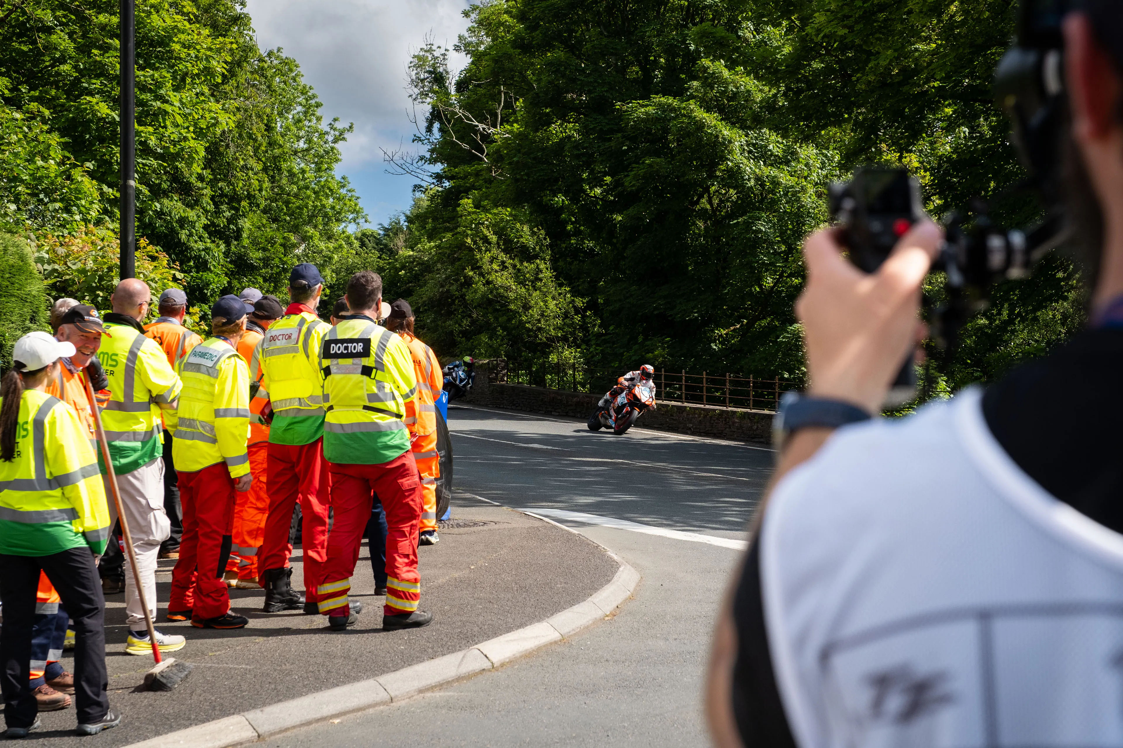 Nico van Loggerenberg filming TT Marshals and MRMS doctors gathered at a bend on the course, with a rider passing during the live TT races