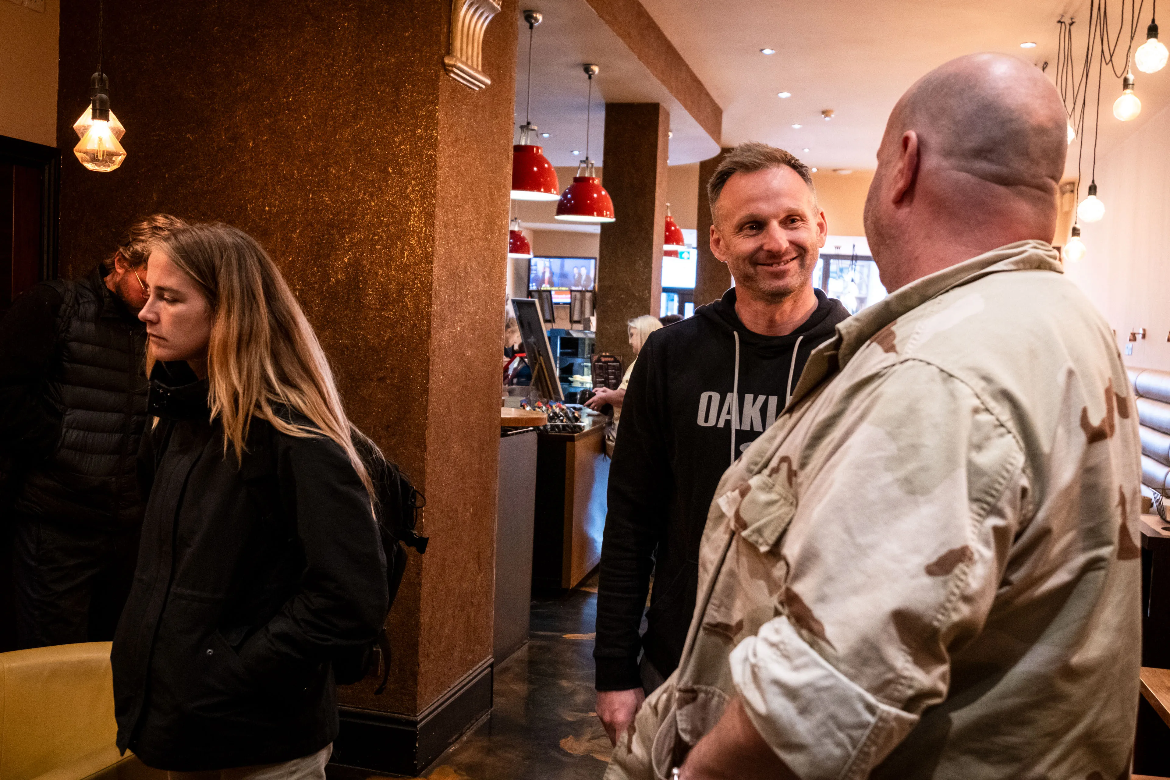 A lively cafe scene with two men chatting and smiling in the foreground, one wearing a black hoodie and the other in a camo shirt. A woman stands to the side, looking down, with warm lighting and modern decor in the background conveying a cozy atmosphere.