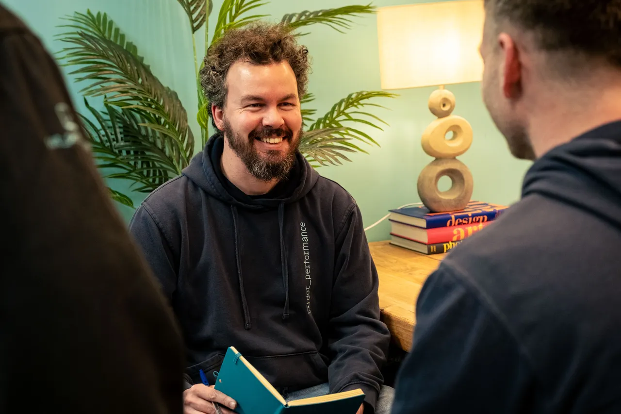 Adrian Crellin in discussion with colleagues at the DotPerformance office in Douglas, Isle of Man, wearing a DotPerformance hoodie and holding a notebook