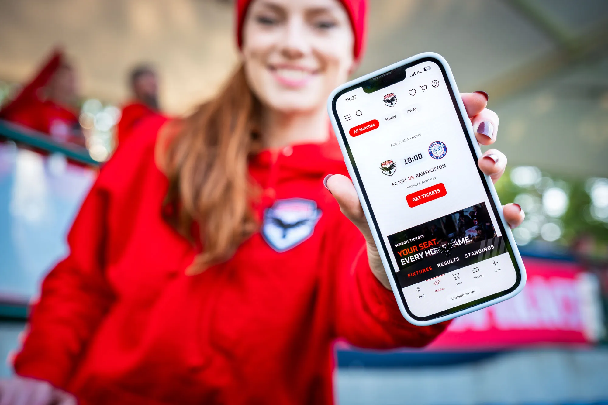 Ella Garvey holding up a phone showing the FCIOM vs Ramsbottom fixture on the updated club website designed by DotPerformance