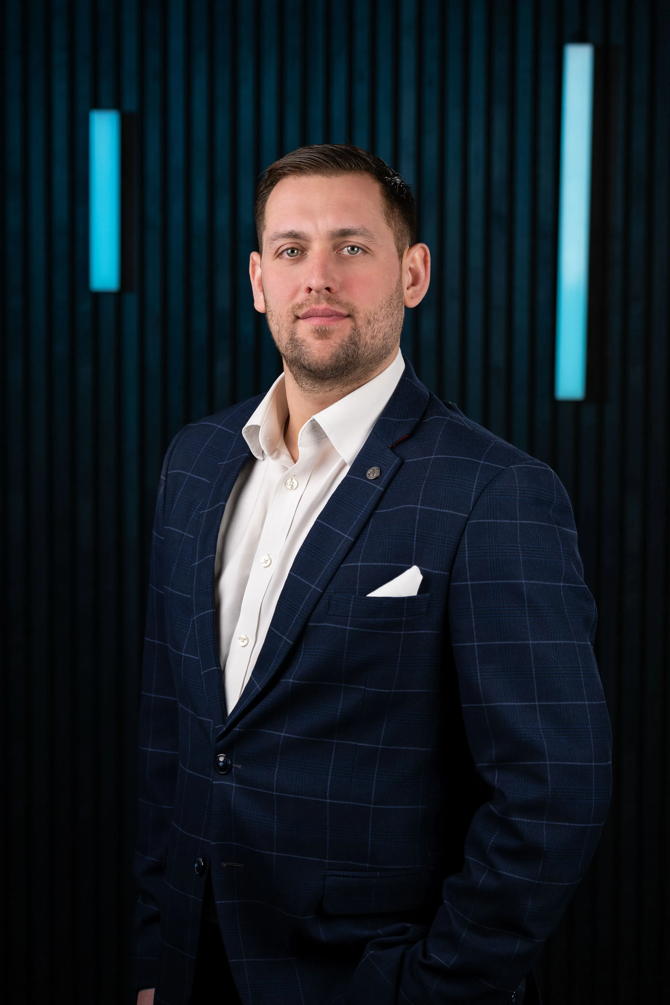 Professional headshot of Brook Longhurst wearing a navy checked blazer and white shirt, against a dark studio backdrop with blue accent lighting