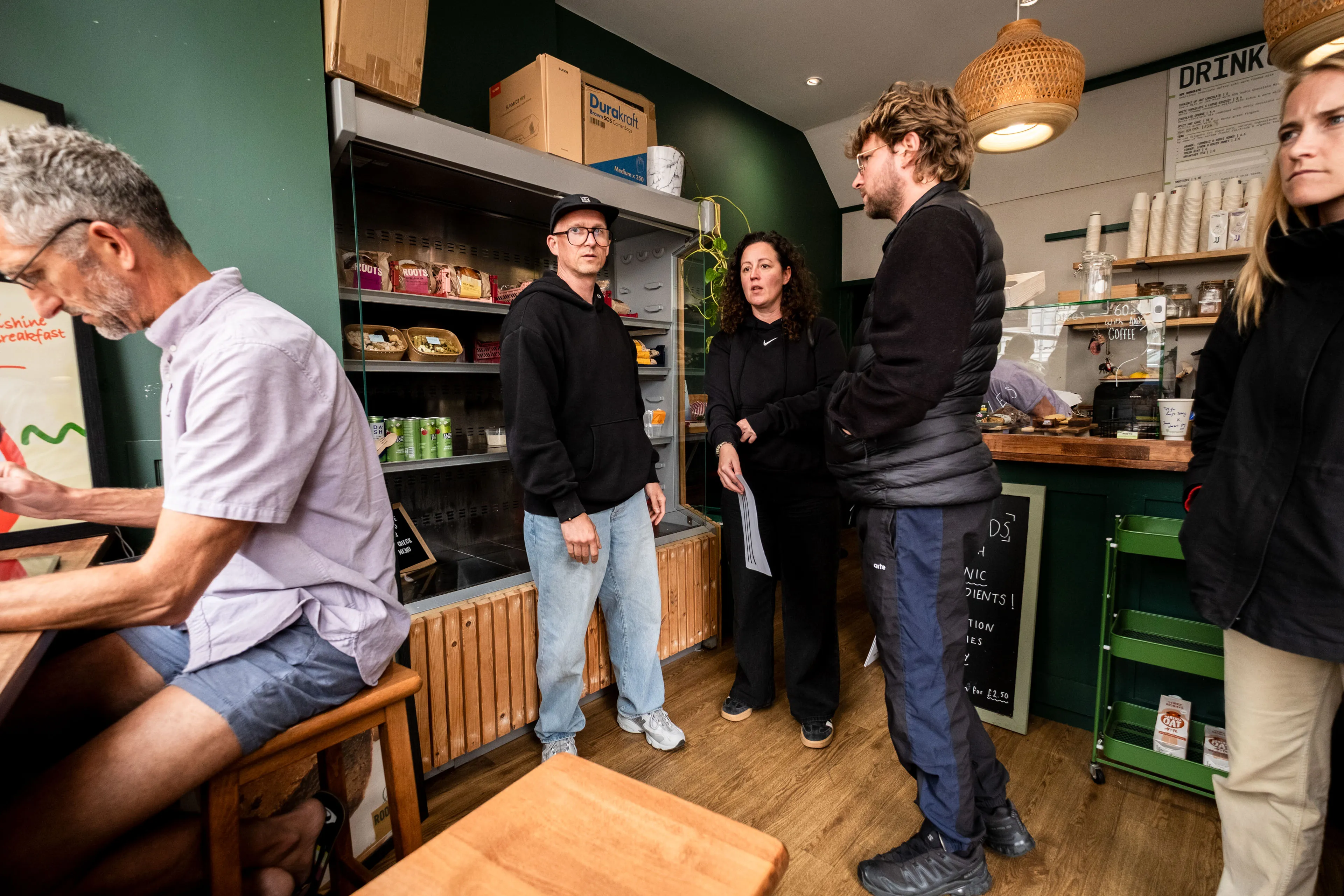 A small cafe with wooden furniture and green walls. Four people stand talking, wearing casual clothes. A menu board and snacks are visible. Cozy and informal atmosphere.