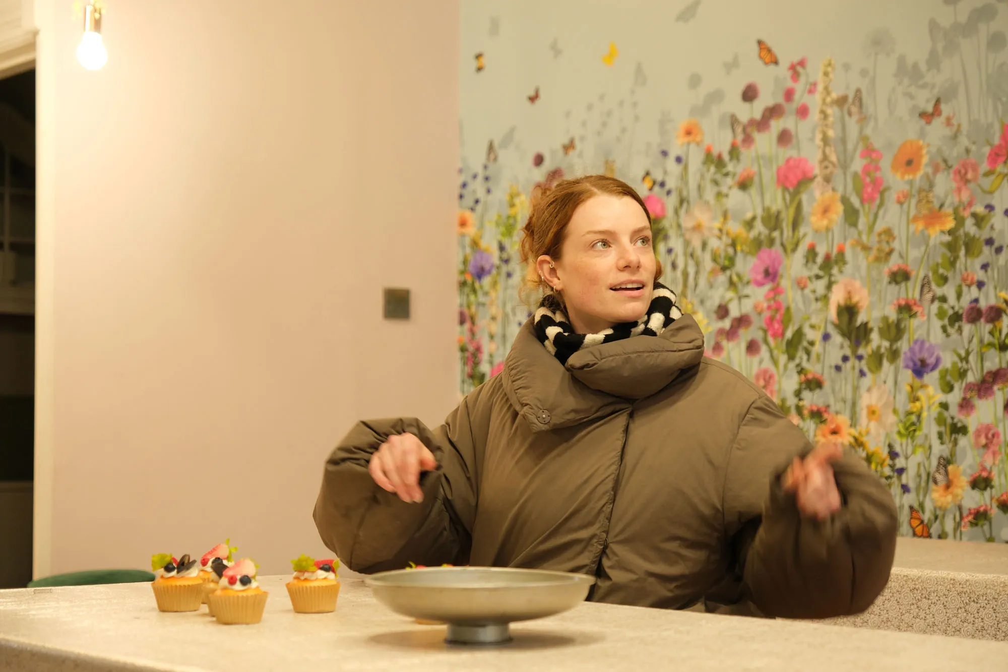 Ella, assistant producer at Dot Performance, mid talking as she gestures to photographer, sat at a table with cupcakes on and flowery wall in background.