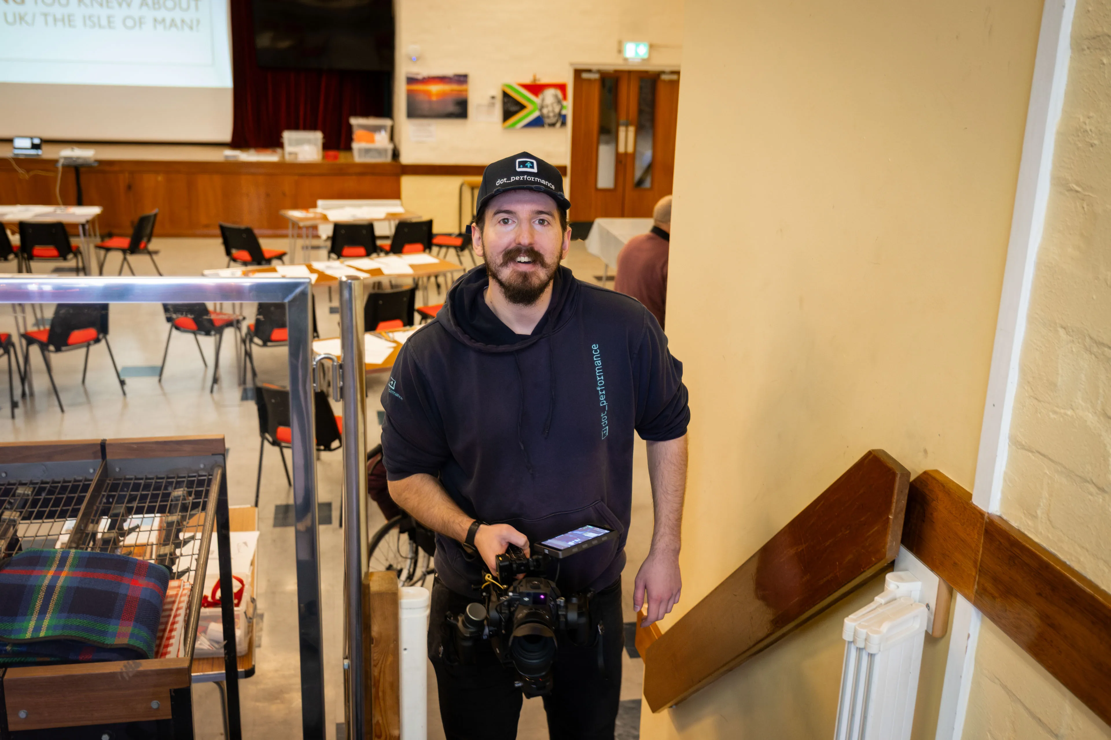 Man with camera stands near staircase in a casual setting, with tables and chairs in the background. The mood is relaxed and informal.