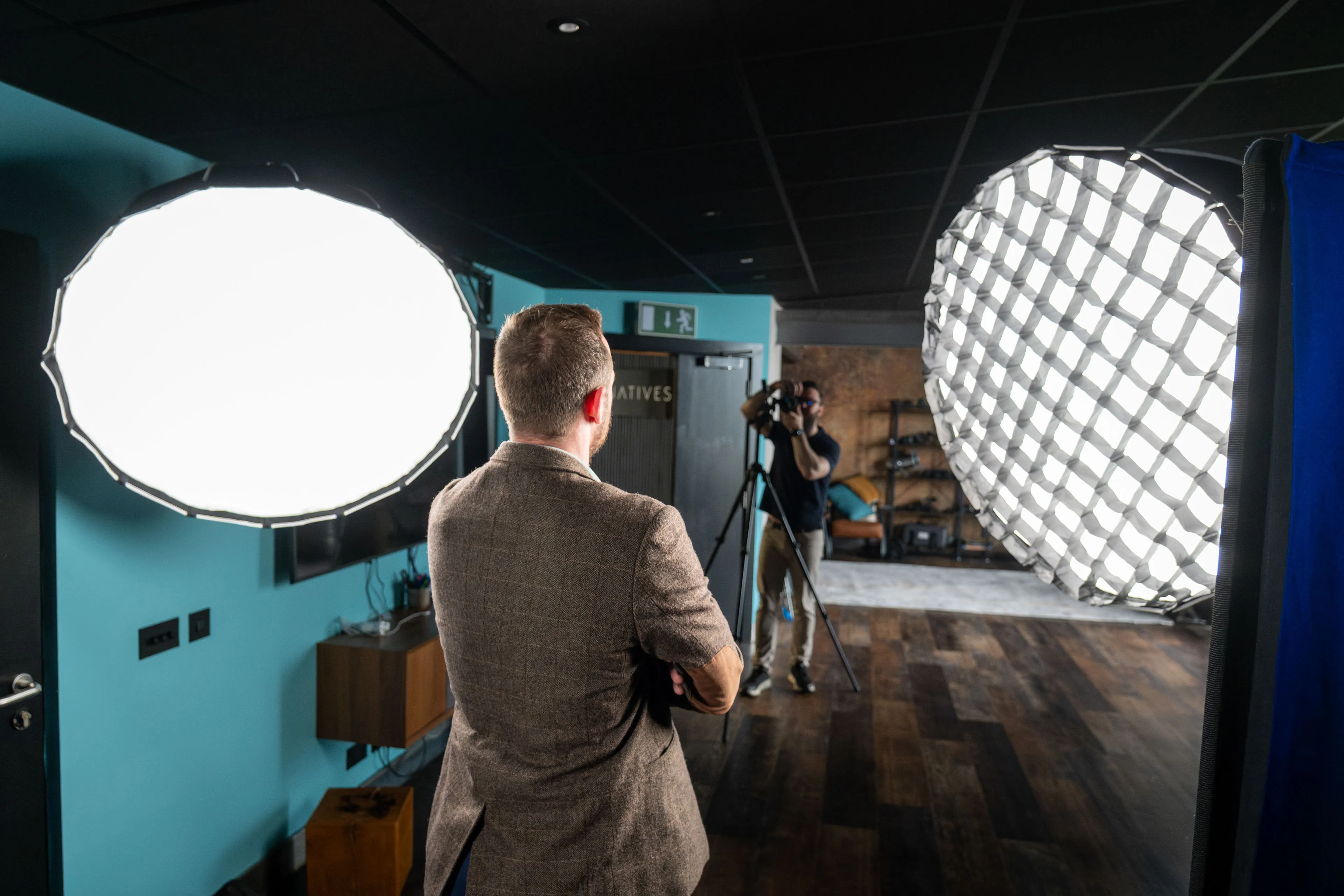 View from behind a headshot subject with arms crossed, facing a softbox light while a photographer works with a camera on a tripod in the background