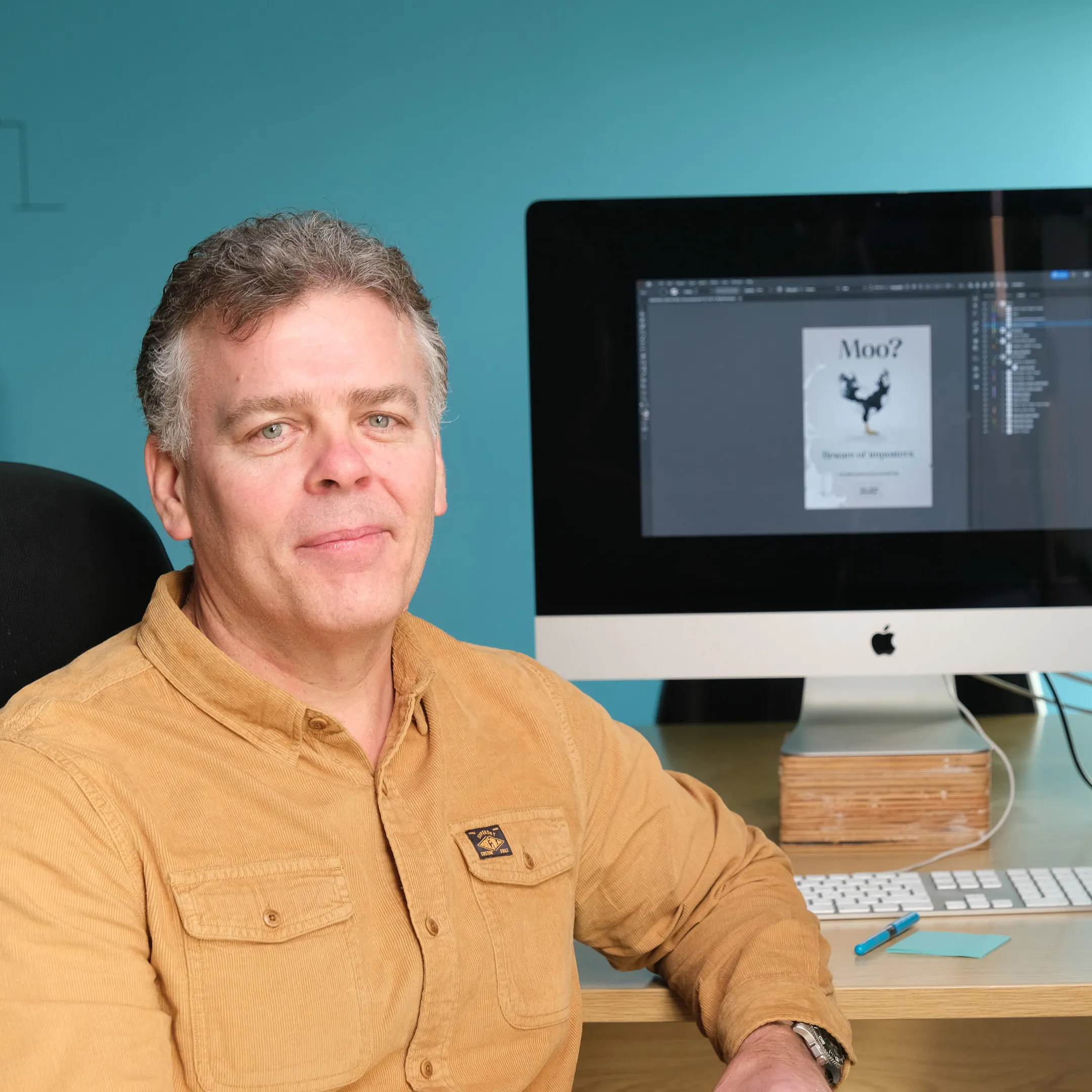 Jason Burton working on a brand design project at his desk at DotPerformance on the Isle of Man, with a graphic design layout open in Adobe Photoshop on an Apple iMac